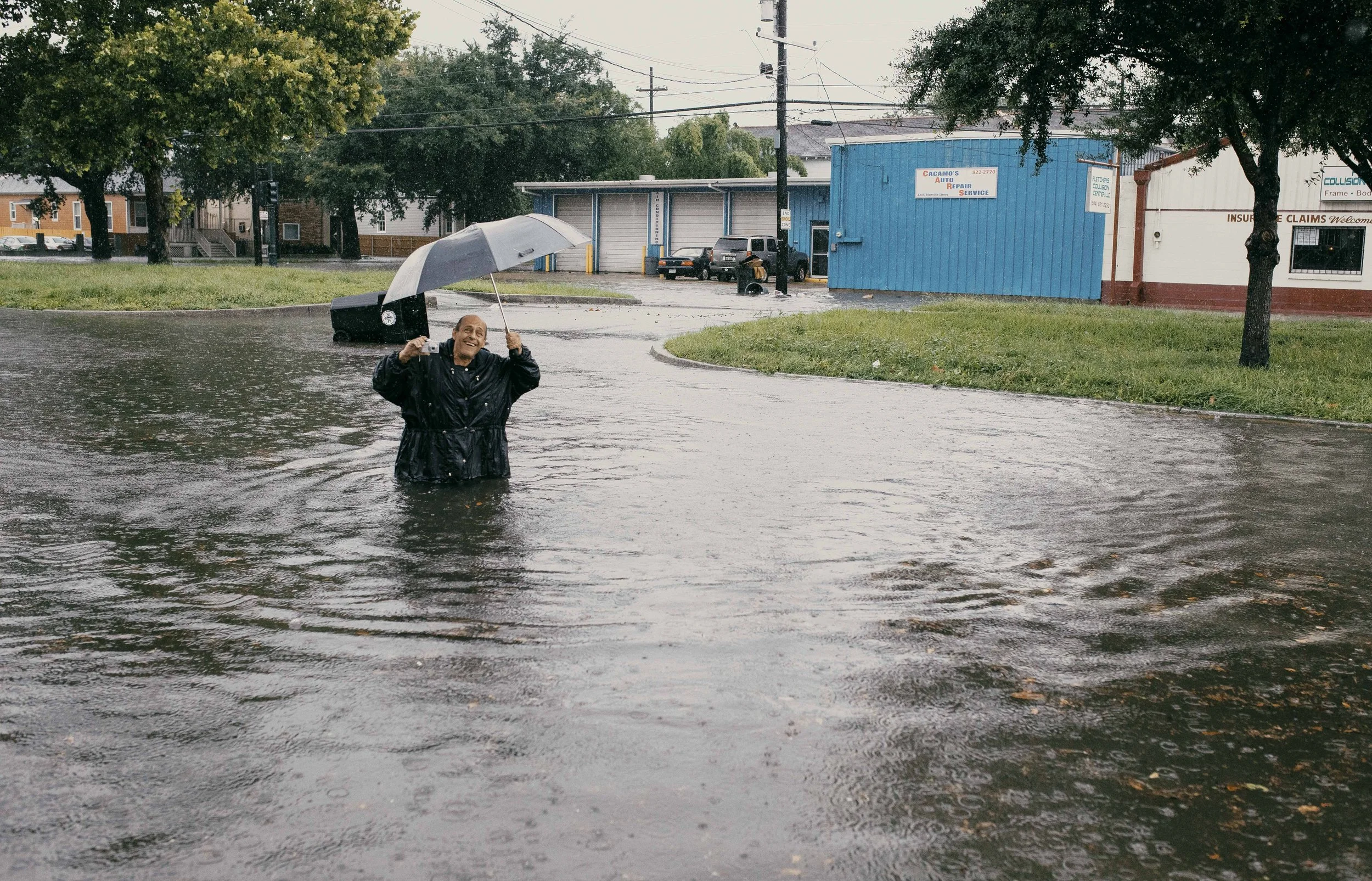 A person standing in a flooded street holding an umbrella and a camera, smiling, with trees, utility poles, and buildings in the background during rainy weather.