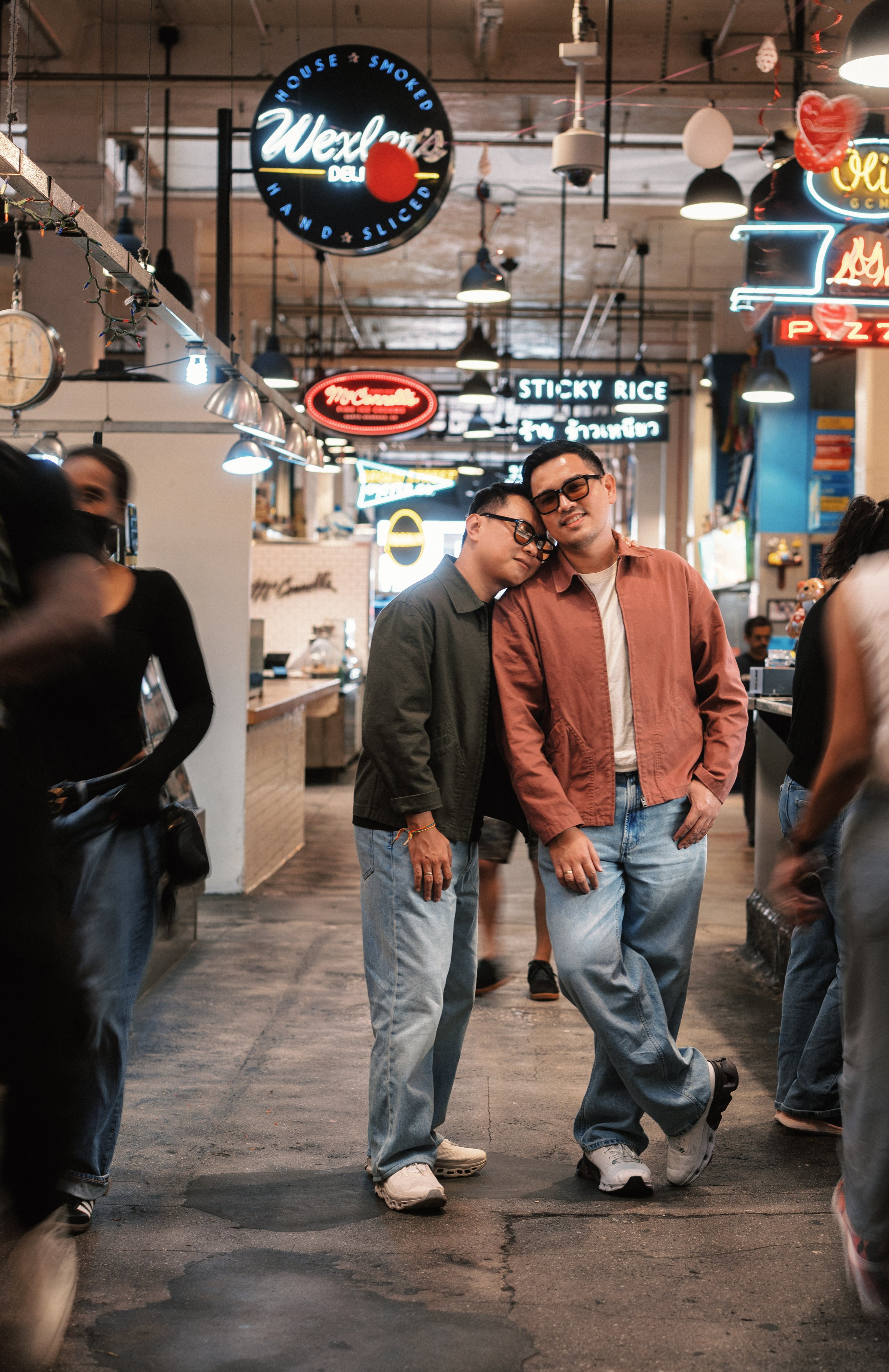 Two people are standing close together with their heads touching, smiling, in a bustling indoor marketplace or food court with neon signs and hanging lights.