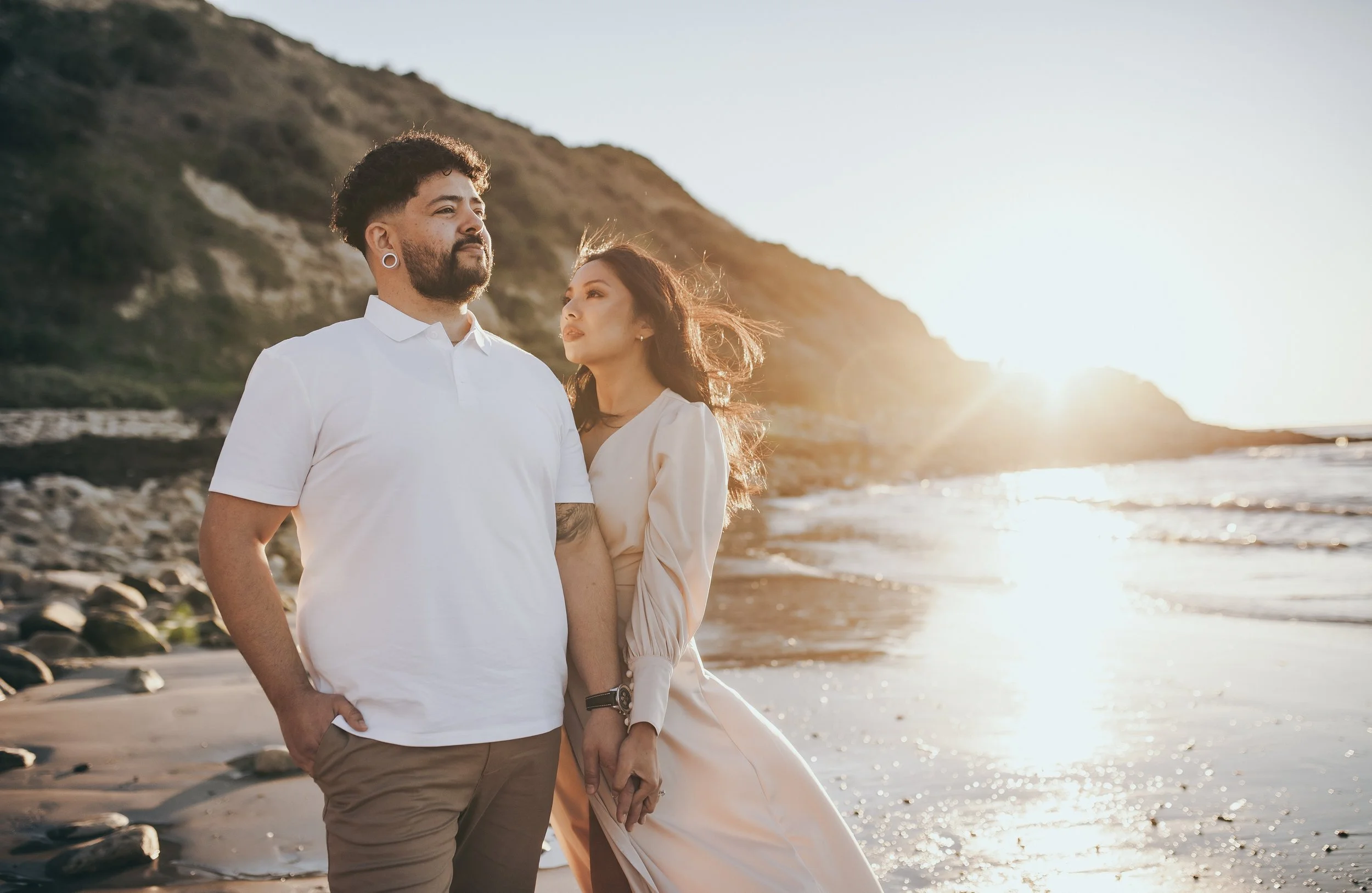 A couple standing on a beach during sunset, with rocky hills in the background, the man with dark curly hair, beard, earrings, wearing a white shirt, and the woman with long dark hair, beige dress, holding hands.