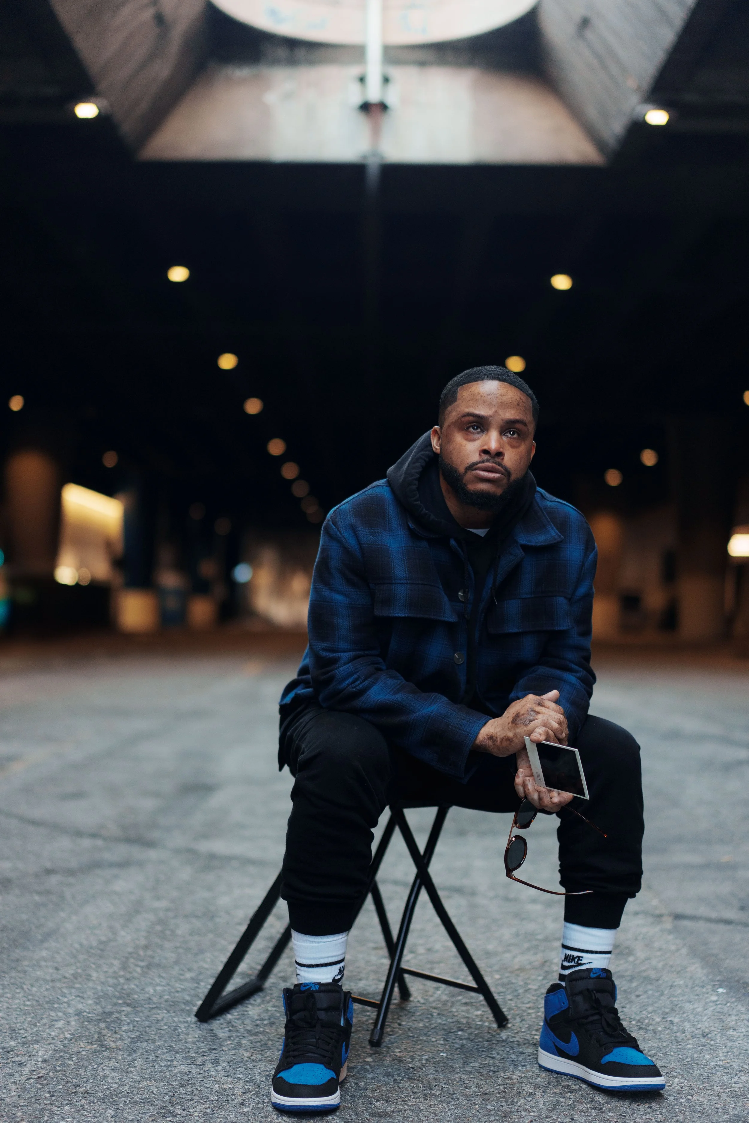 A man sitting on a small stool outdoors on a city street at night, holding a smartphone in one hand, with sunglasses hanging from his other hand, wearing a blue plaid jacket, black pants, and black and blue sneakers, with a dark background and blurre