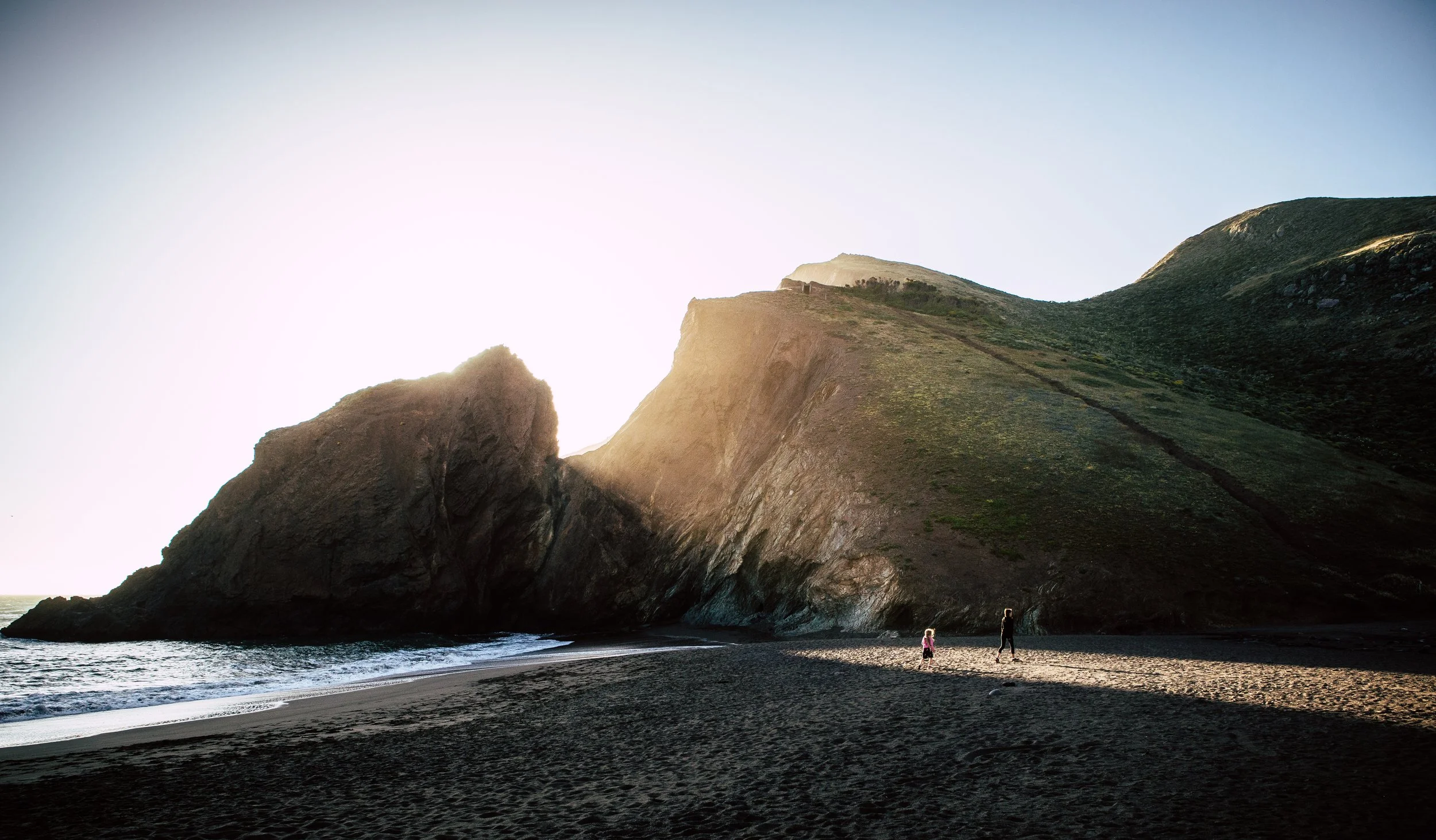 People walking along a sandy beach with large rocky cliffs and green hills in the background, sunlight shining over the landscape.