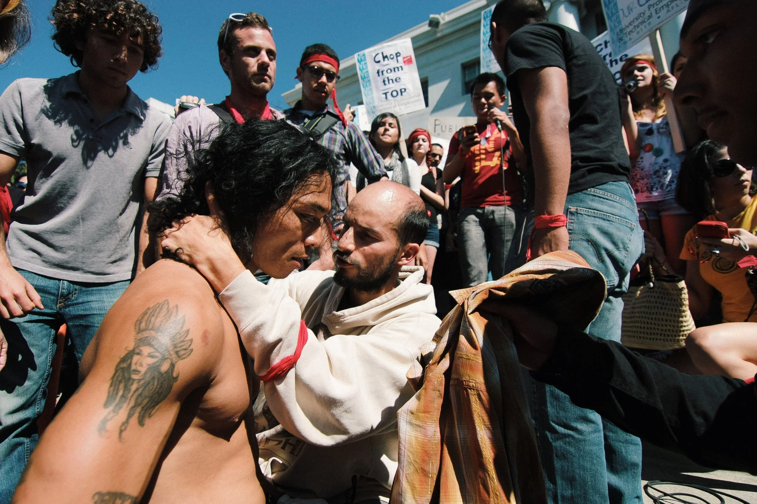 A diverse group of protesters gather outdoors, some holding signs, engaging in a demonstration during daytime.