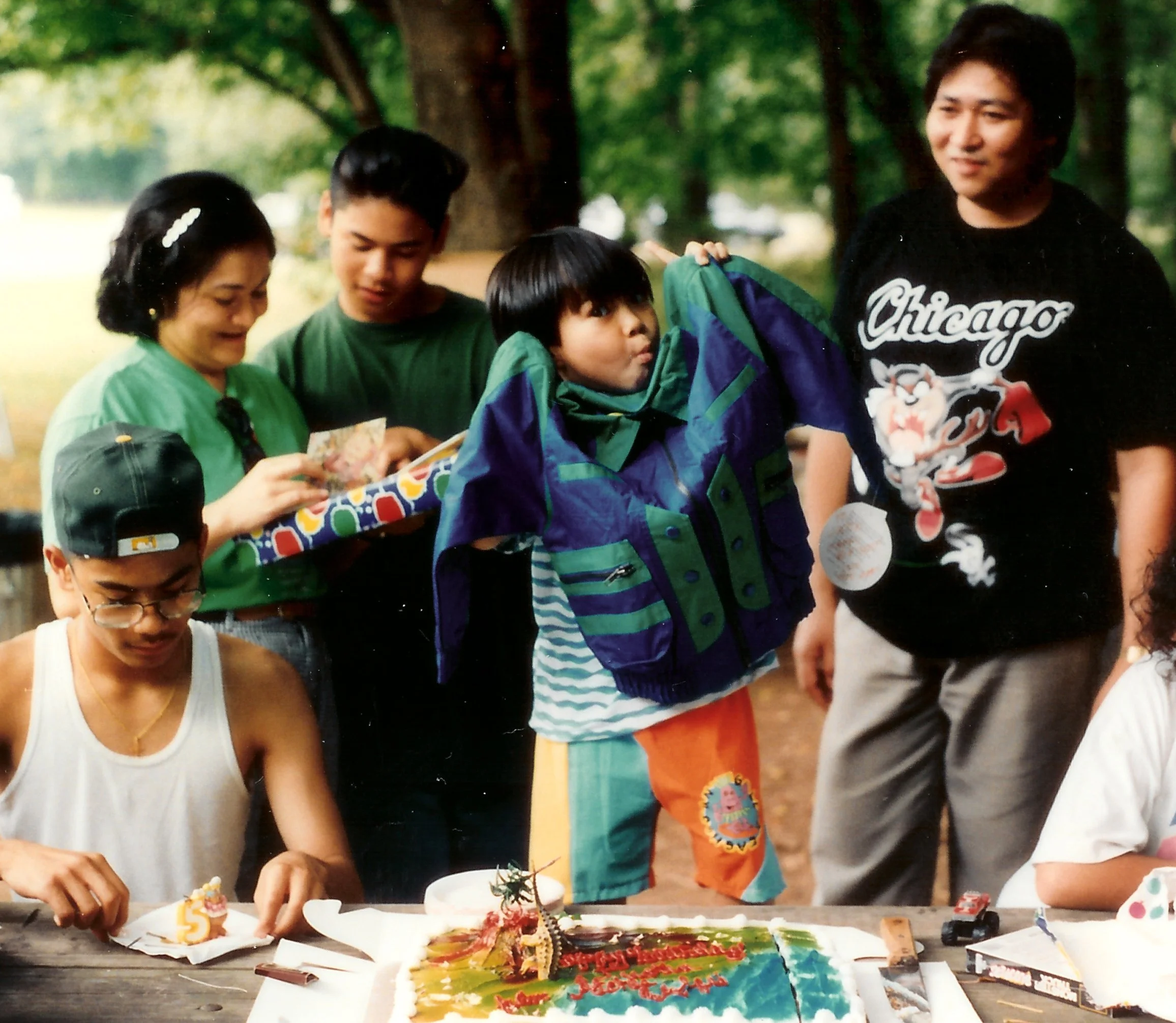 A birthday celebration outdoors with children and adults gathered around a table with birthday cakes, party decorations, and presents, in a park or garden setting.