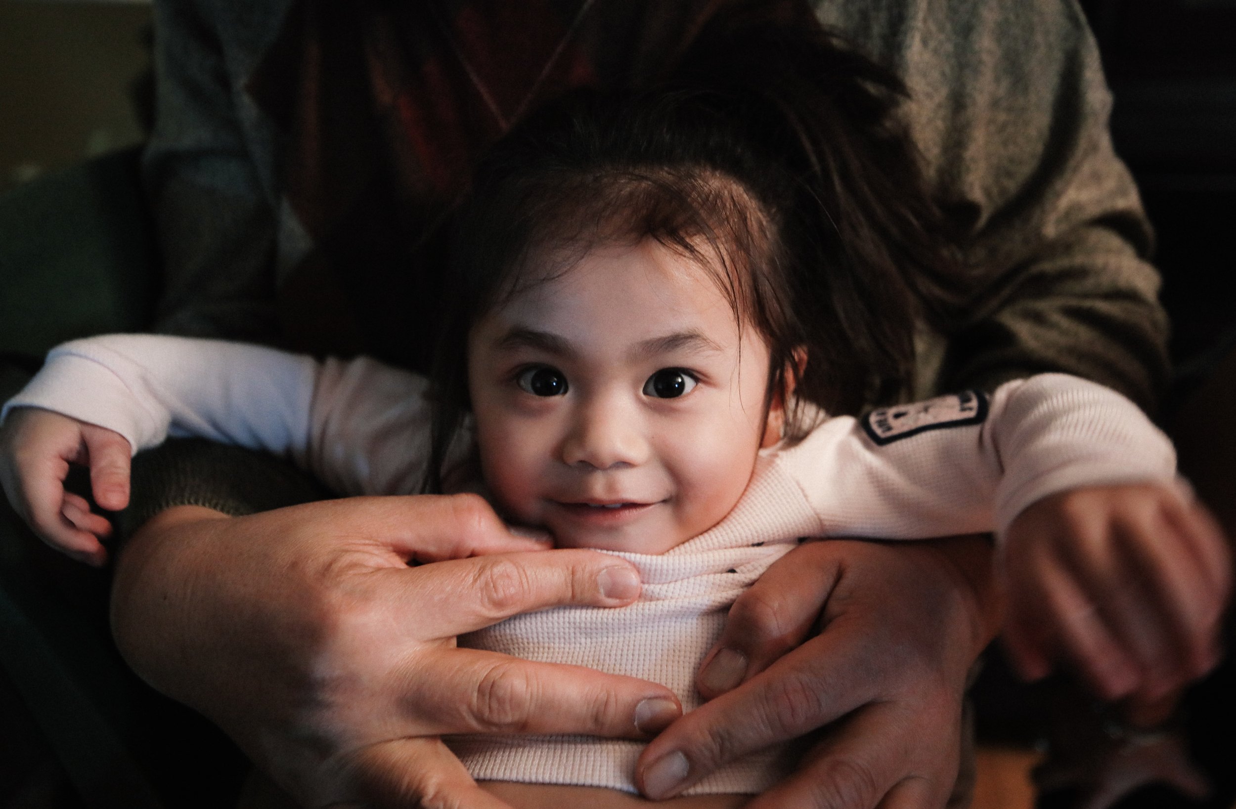 A young girl with big, expressive eyes lying on her stomach, being gently held by an adult. The girl is wearing a light-colored shirt. The setting appears to be indoors with dark background lighting.