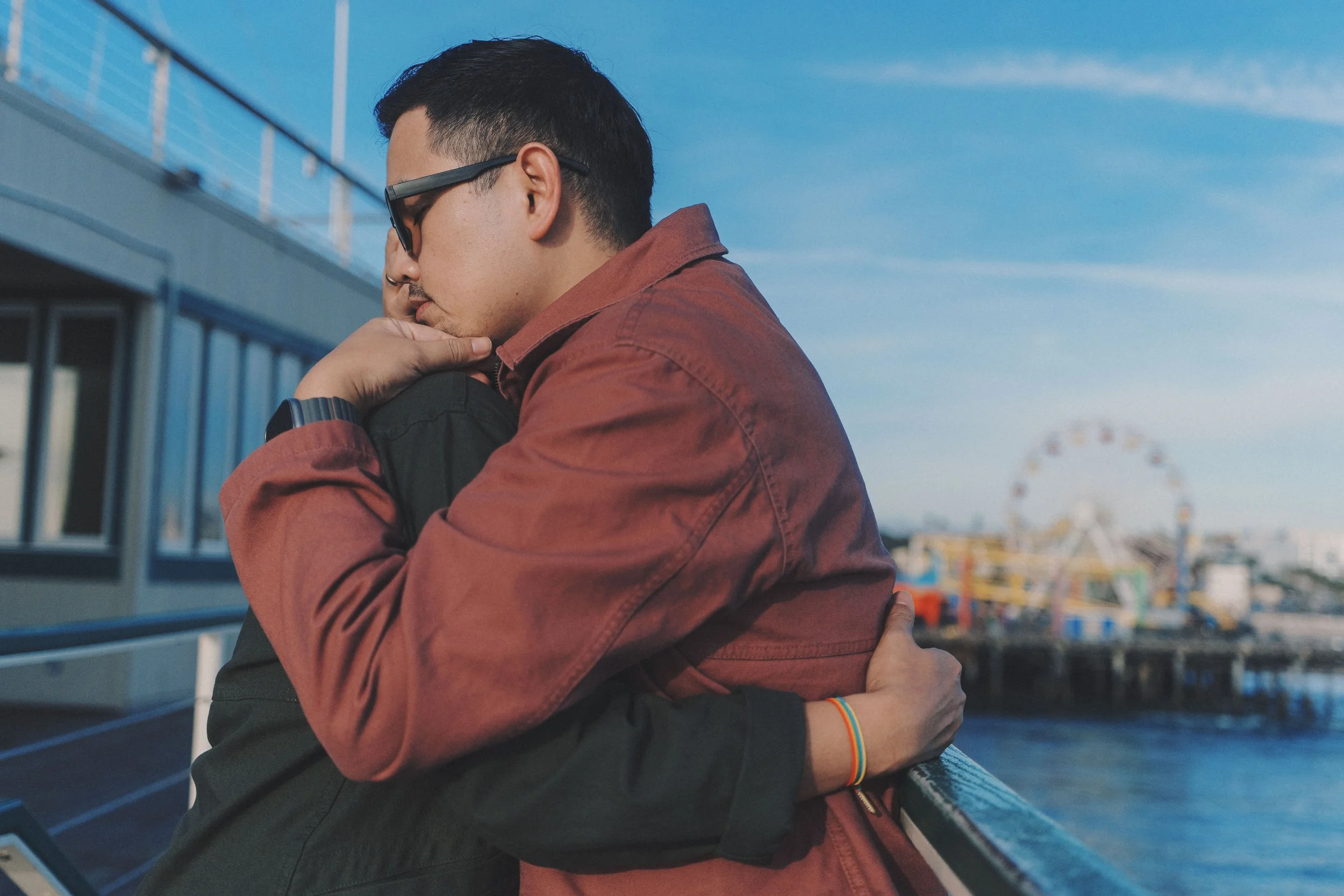 Two people hugging on a deck overlooking the ocean with a Ferris wheel in the background on a clear day.
