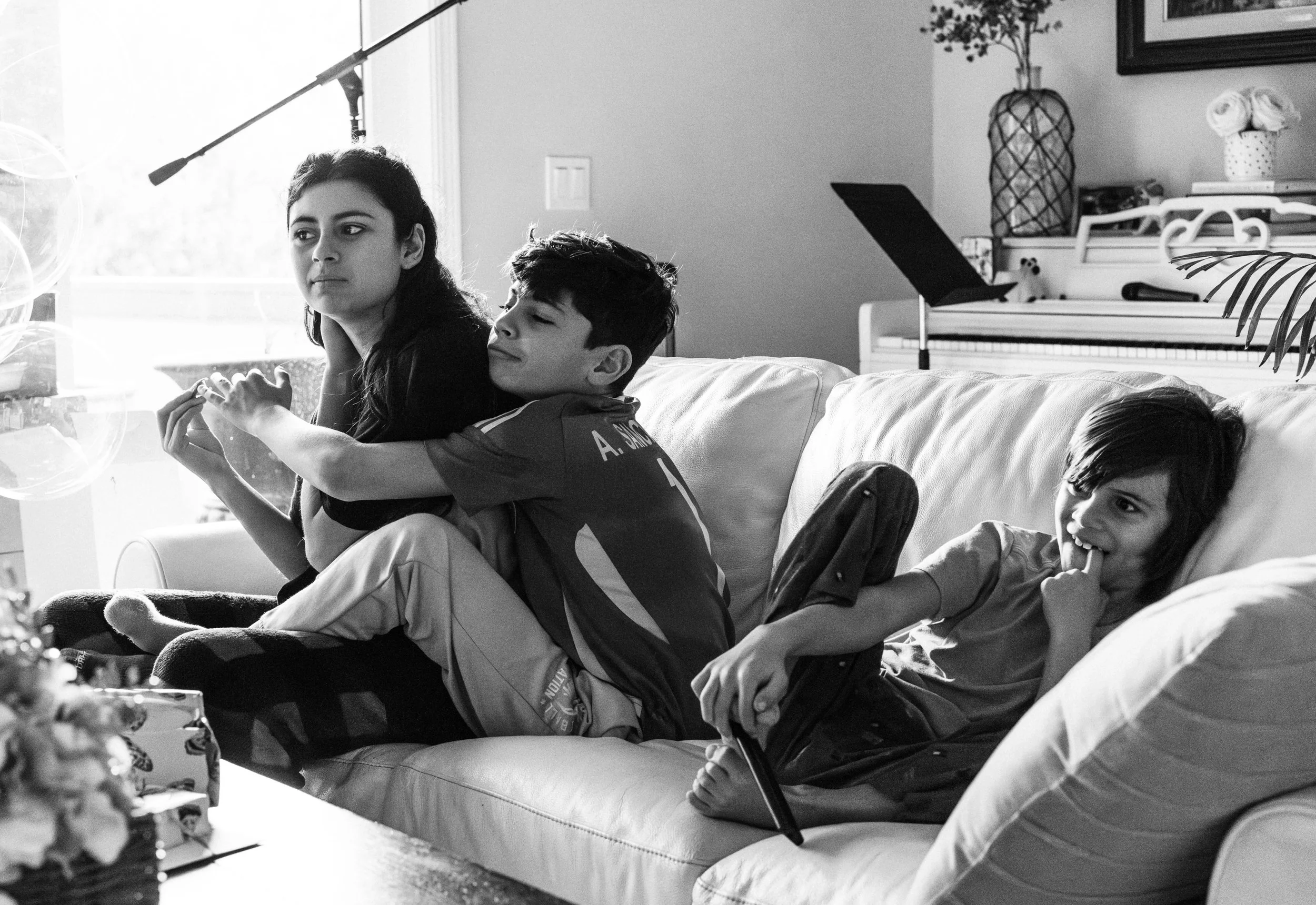 Two kids sitting on a sofa in a living room, one girl hugging another boy, with a piano and decorative items in the background.