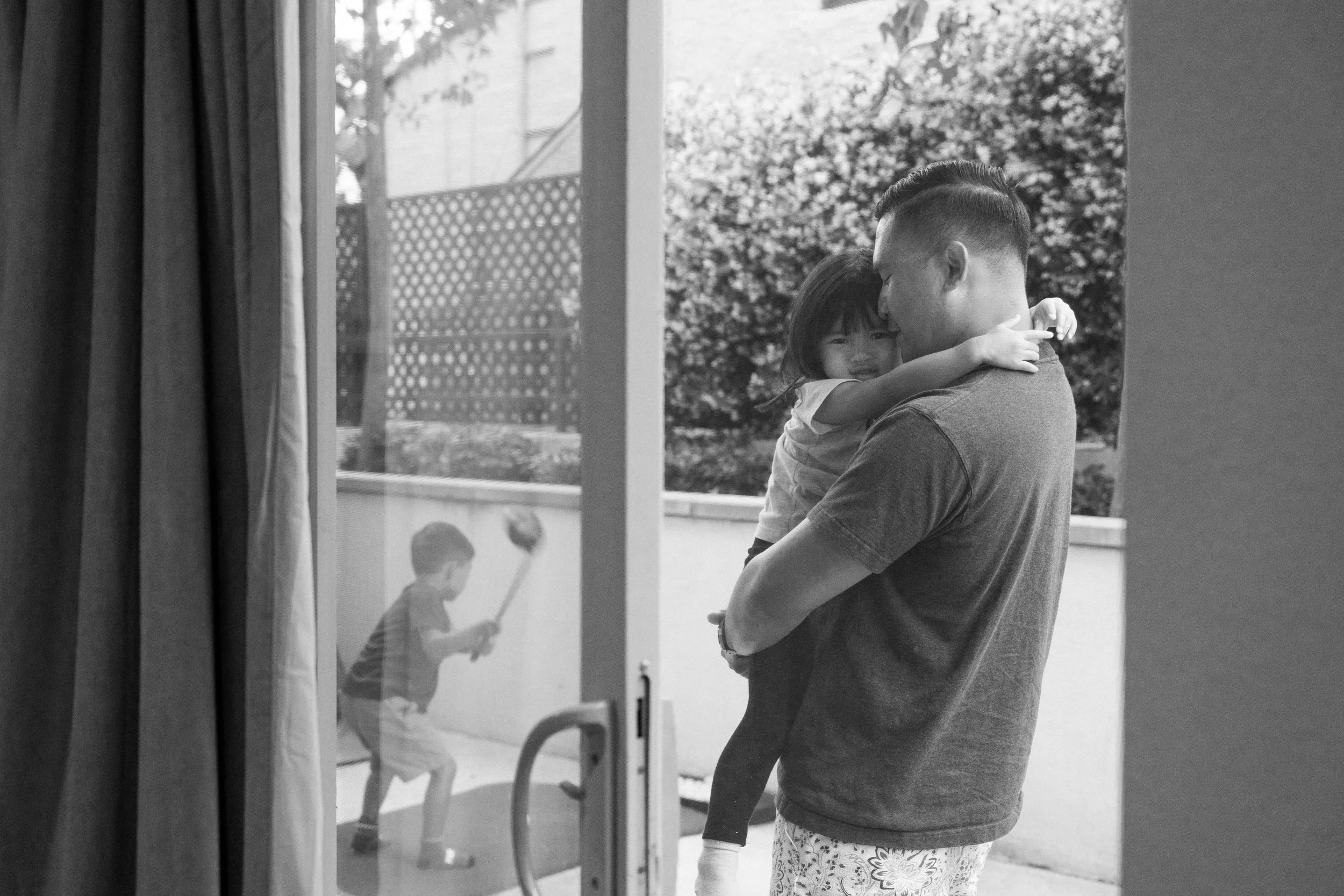 A man holding a young girl close, both with foreheads touching, inside a house looking out a window at children playing outside on a playground.