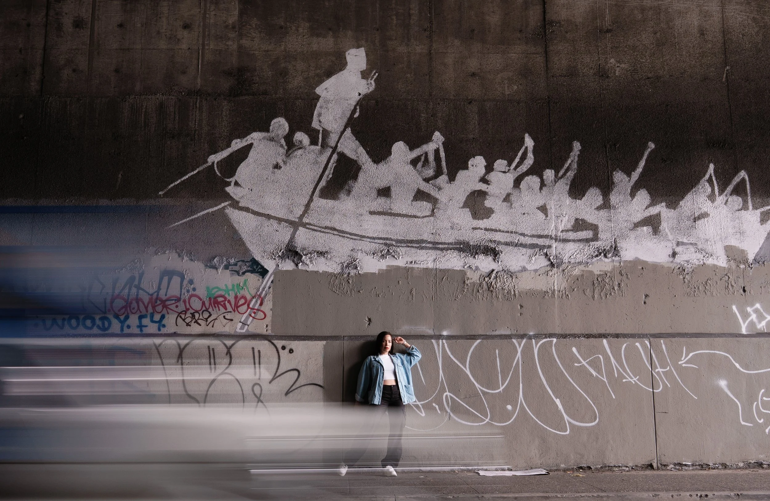 A young woman in casual clothing standing and leaning against a graffiti-covered wall with a white graffiti artwork of a group of people rowing a boat, with some graffiti tags below.
