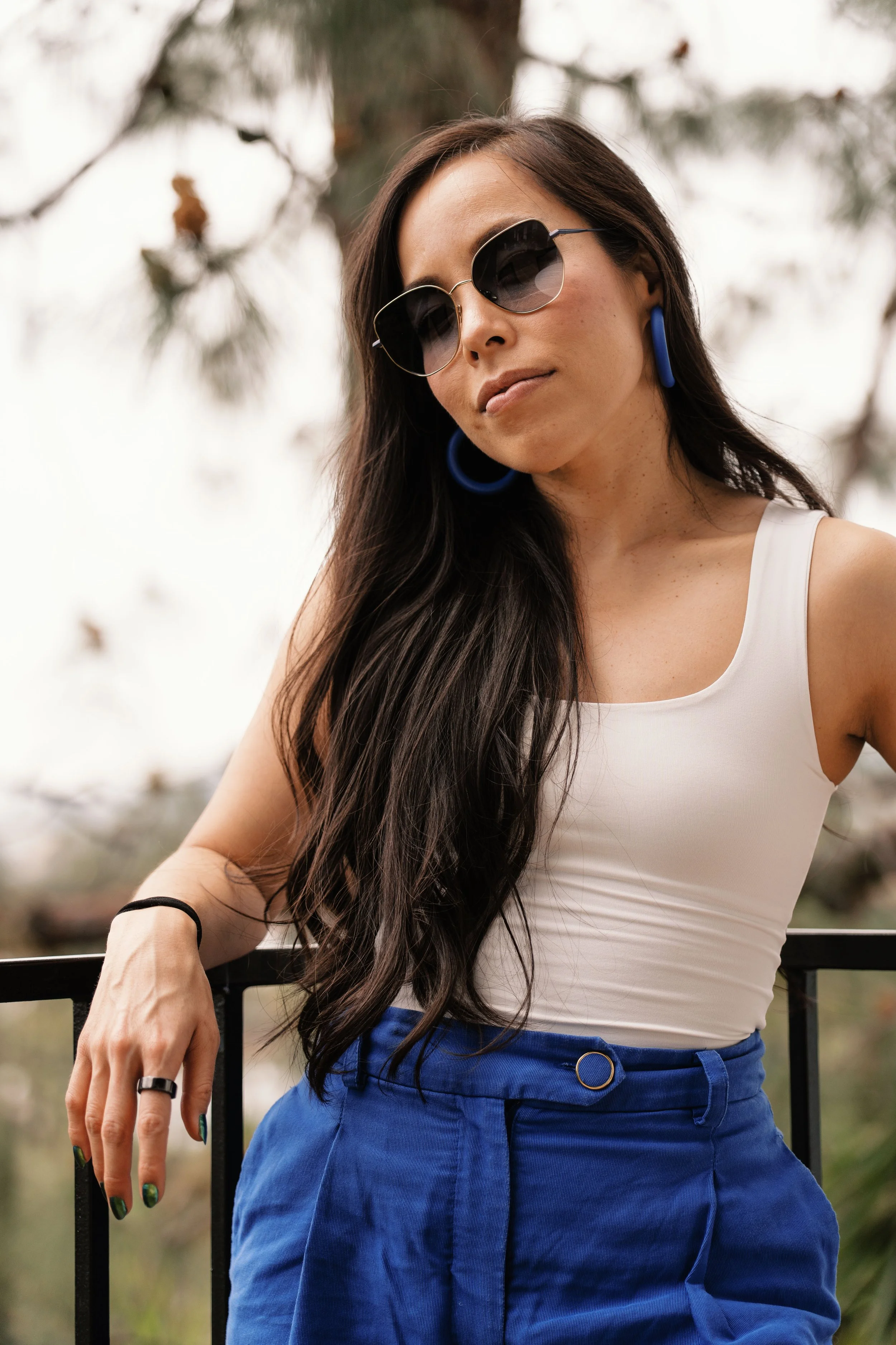 A woman with long dark hair wearing sunglasses, blue earrings, a white tank top, and blue pants, leaning on a black railing outdoors with trees in the background.