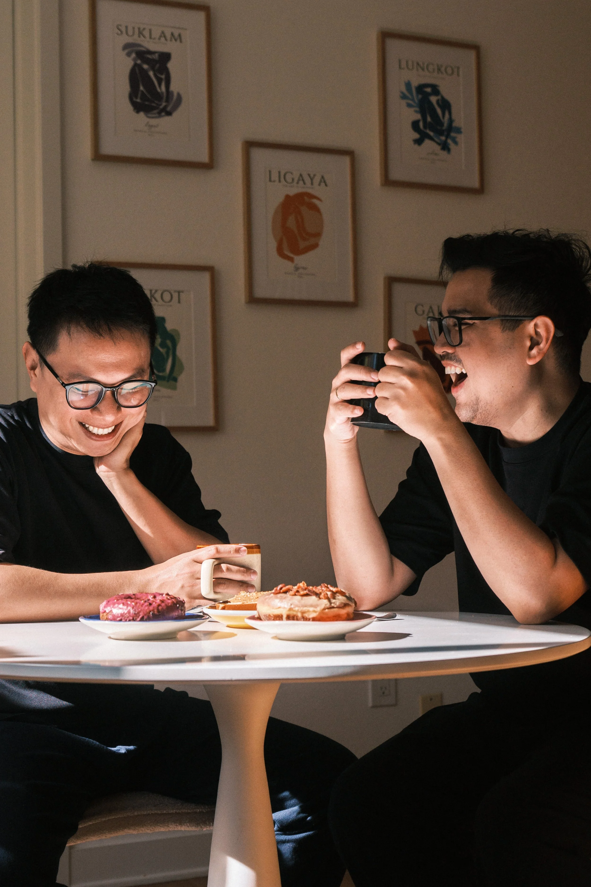 Two men sitting at a table with donuts and coffee, smiling and laughing, in a room with framed artwork on the wall.