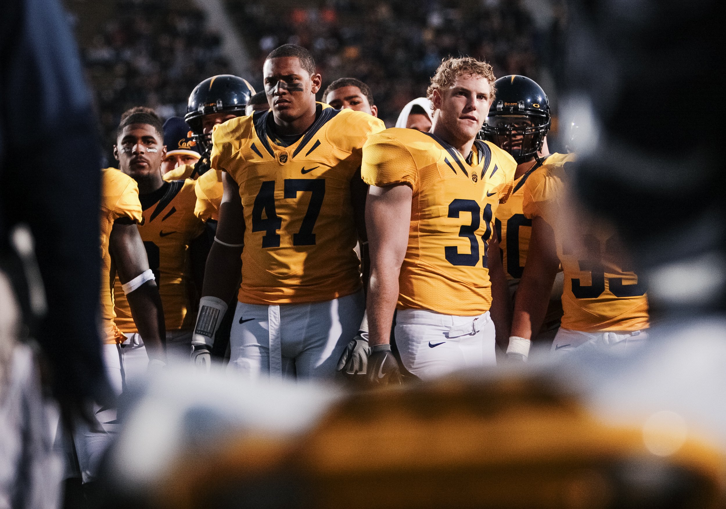 American football players in yellow jerseys standing on the field during a game, viewed through a blurred foreground.