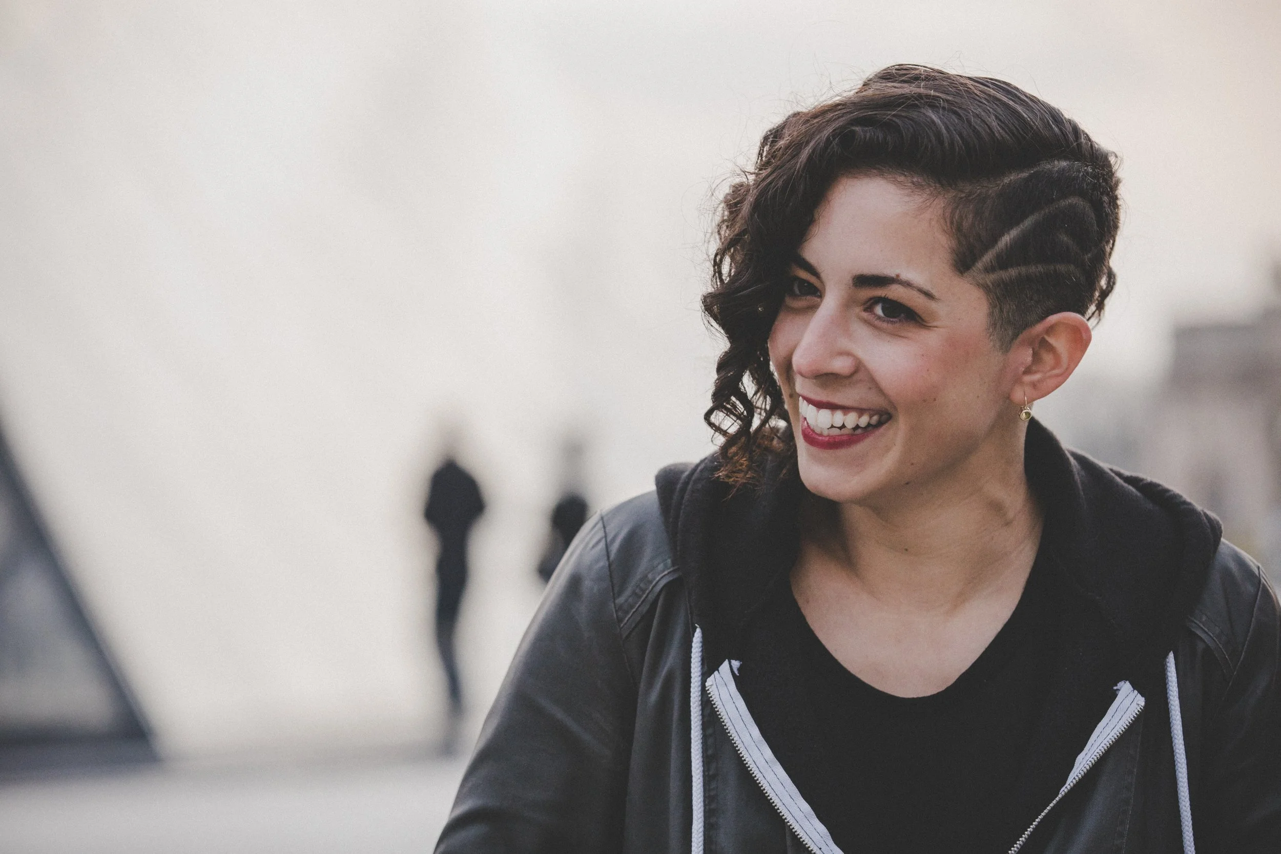 A woman with short dark hair and a side undercut, smiling outdoors on a cloudy day, wearing a black jacket with a striped zipper.
