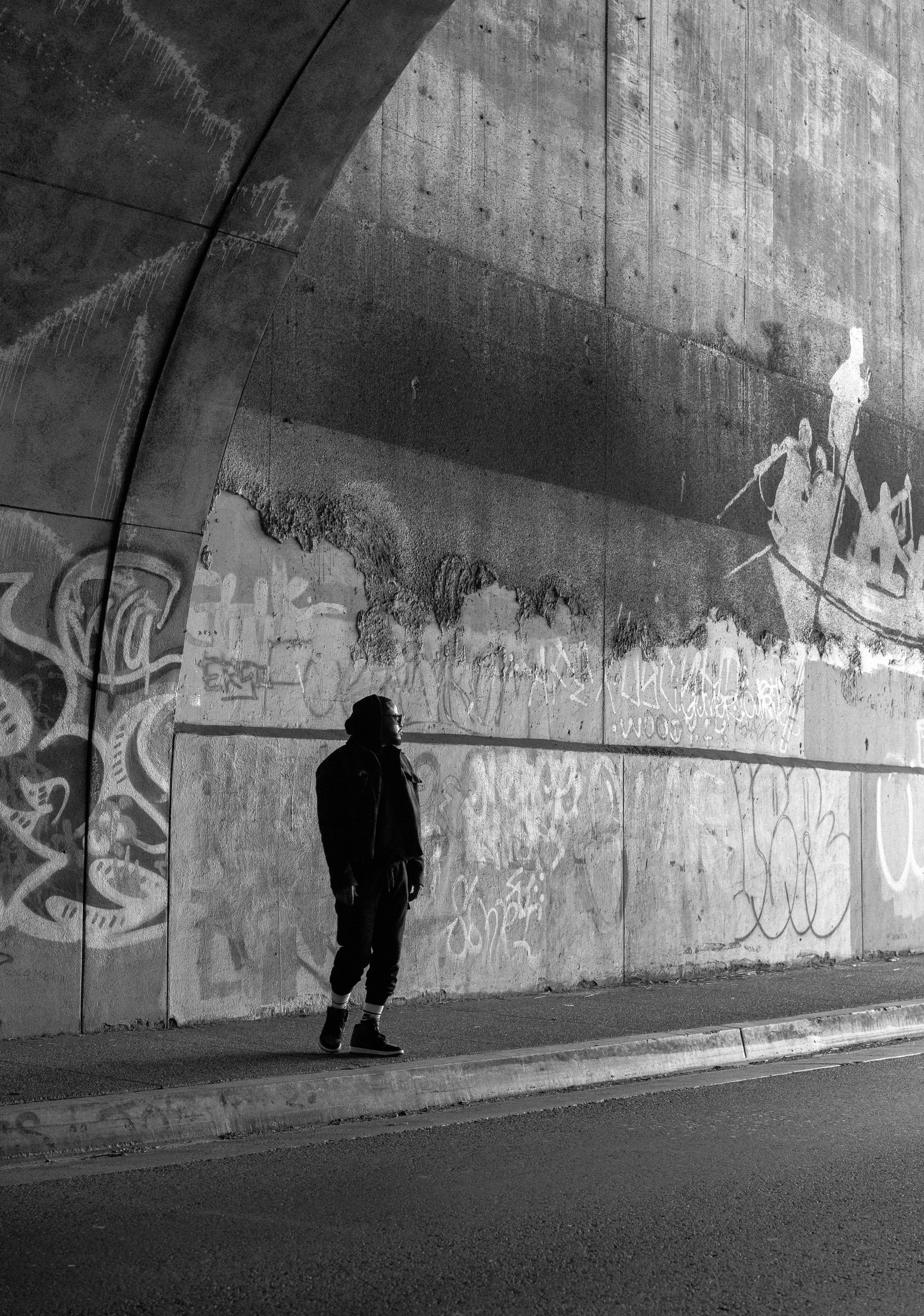 A person standing on the sidewalk under a large concrete bridge with graffiti, with a shadow of the bridge cast on the wall behind them.