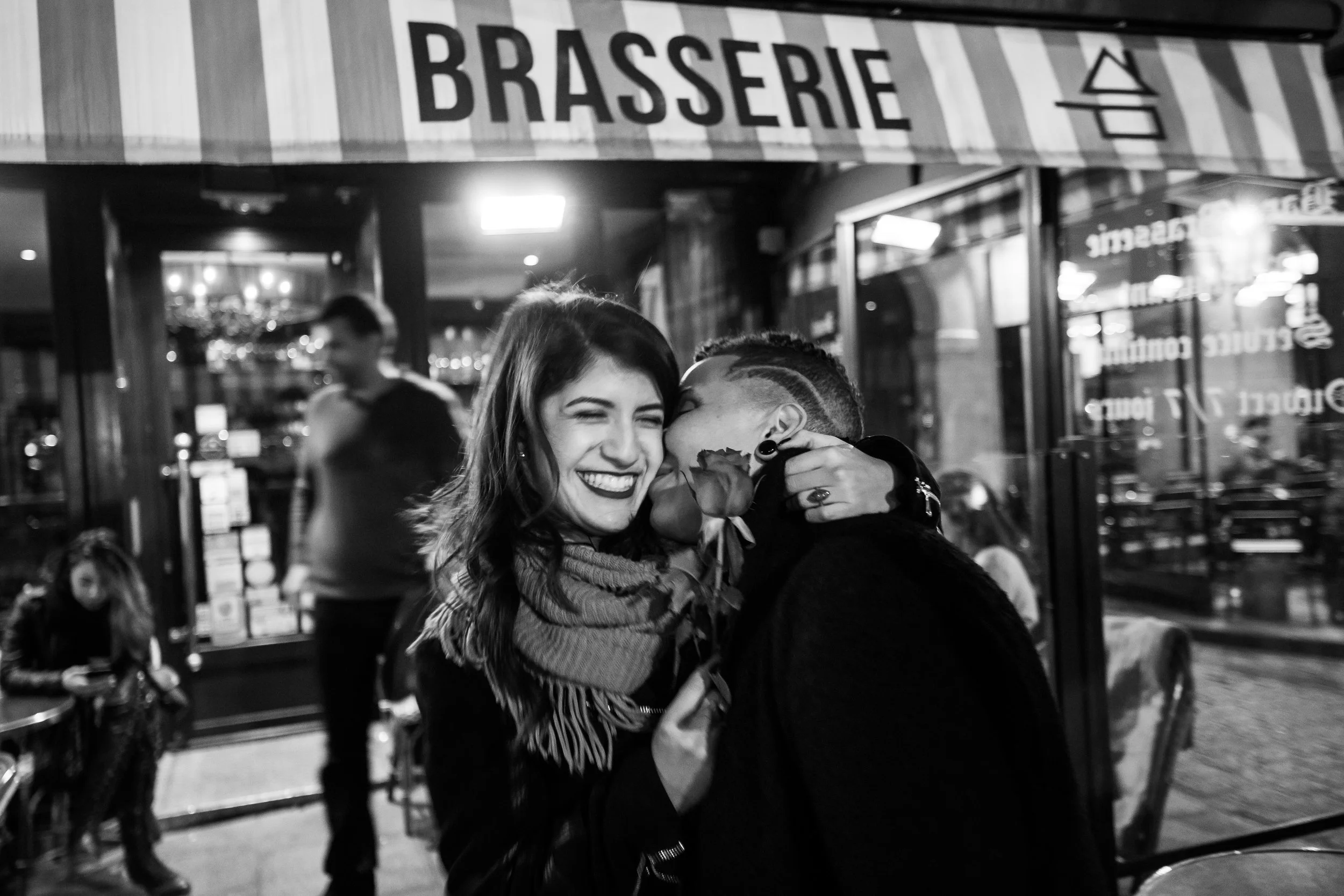 Two people hugging and smiling inside a restaurant or café, with a sign reading 'BRASSERIE' visible above. One person has long hair and the other has a short hairstyle with a shaved side.