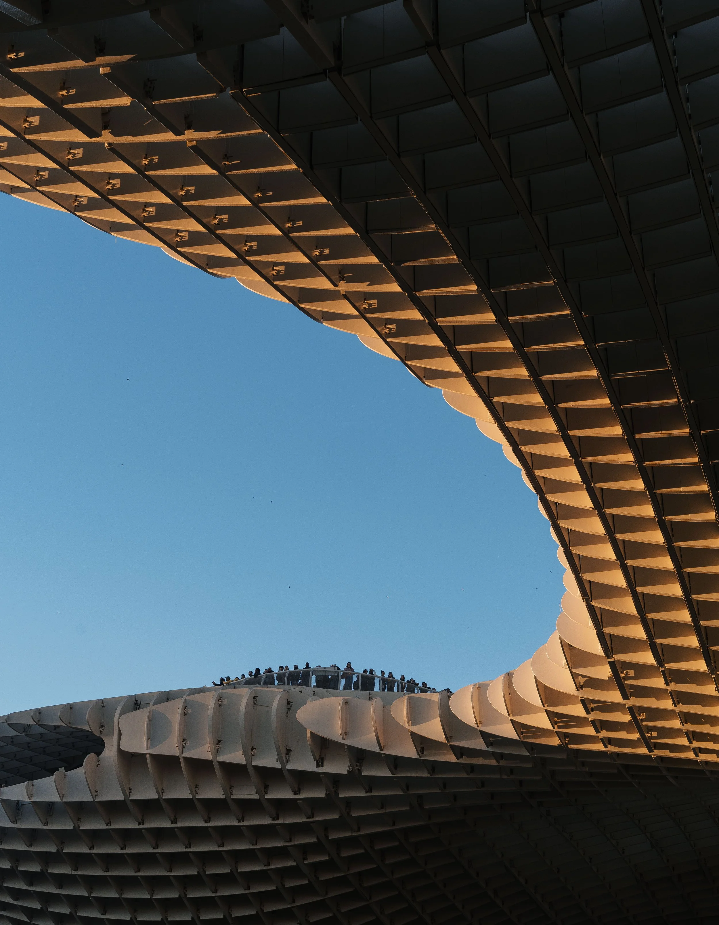 Close-up of a modern building with curved, overlapping panels forming an architectural design against a clear blue sky. People are visible on a balcony at the lower part of the image.