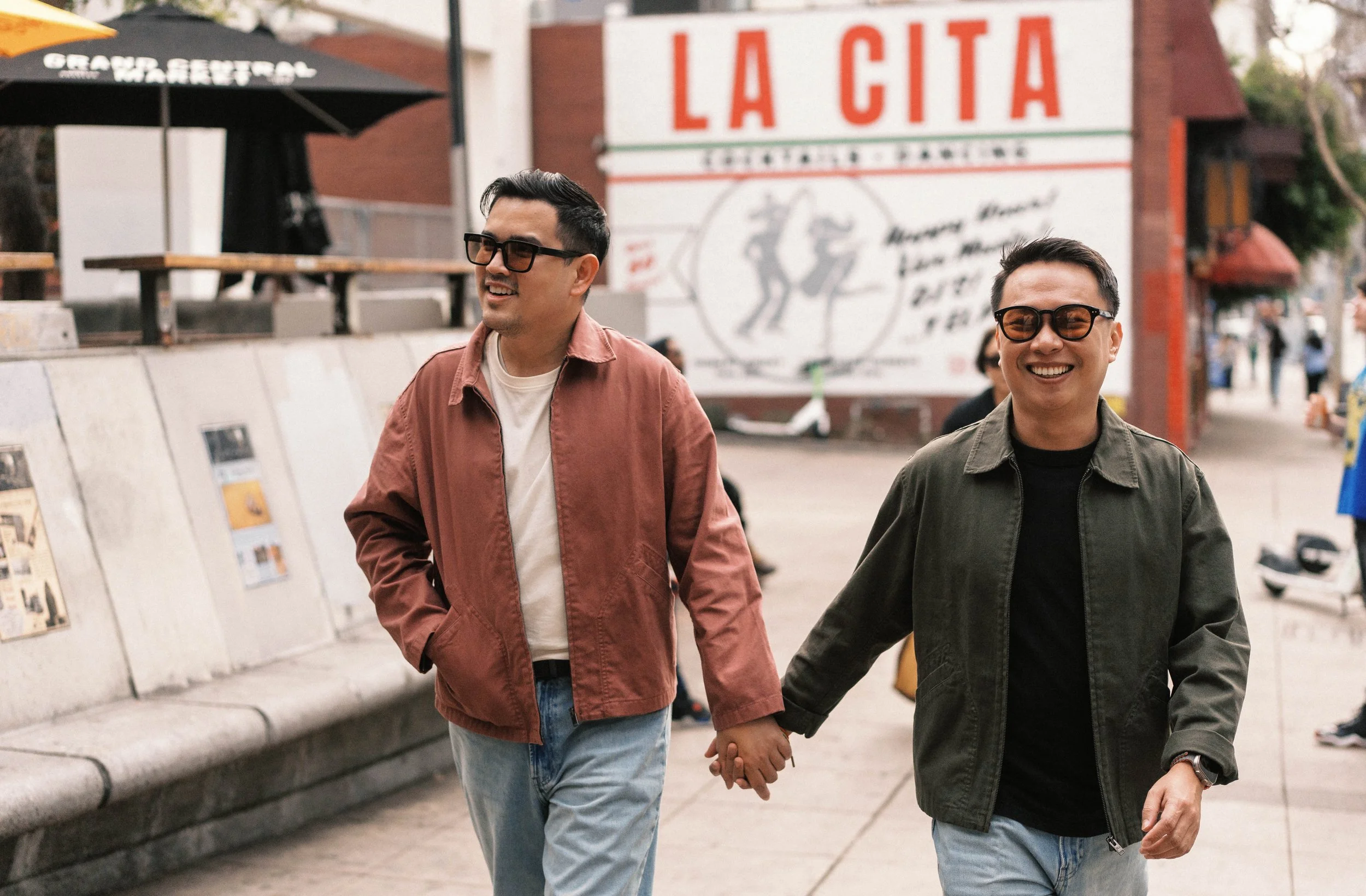Two men walking hand in hand outdoors in front of a sign that reads 'La GITA' with other smaller texts and images, smiling and wearing glasses and casual clothes.