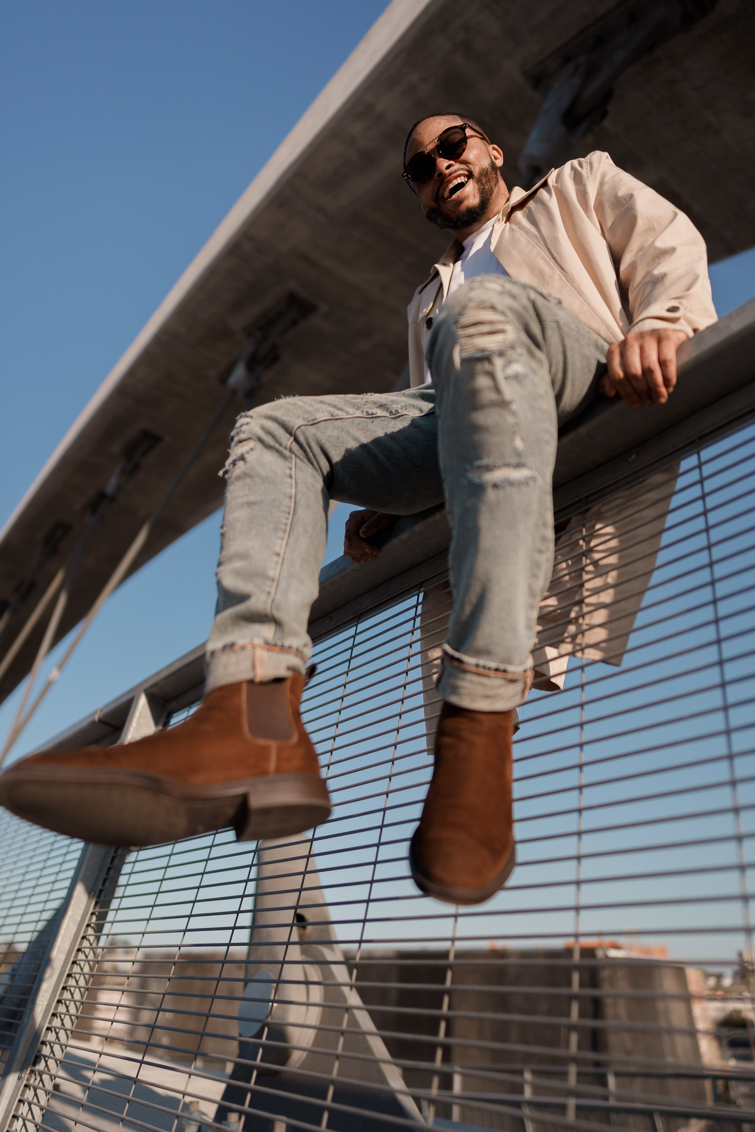 A man with a beard wearing sunglasses, a beige jacket, a white shirt, ripped light-wash jeans, and tan boots sitting on a metal railing under a bridge, smiling and looking down at the camera against a blue sky.