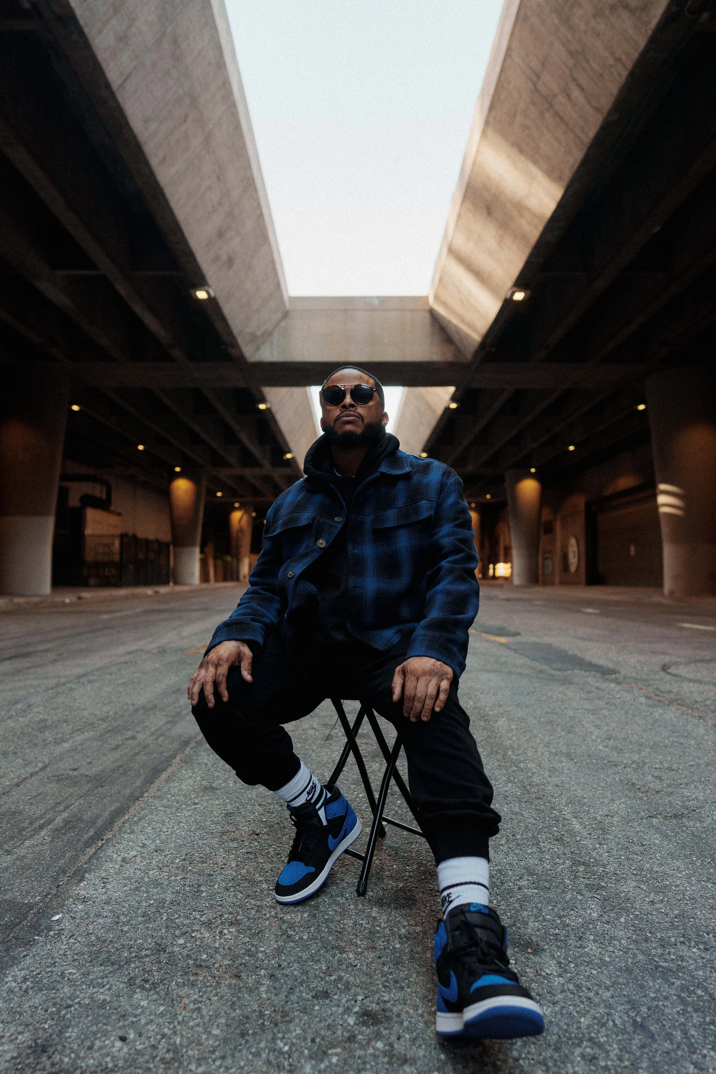 A man wearing sunglasses, a blue plaid jacket, black pants, and sneakers is sitting on a foldable chair in the middle of an empty parking lot under an overpass, with a large skylight overhead.