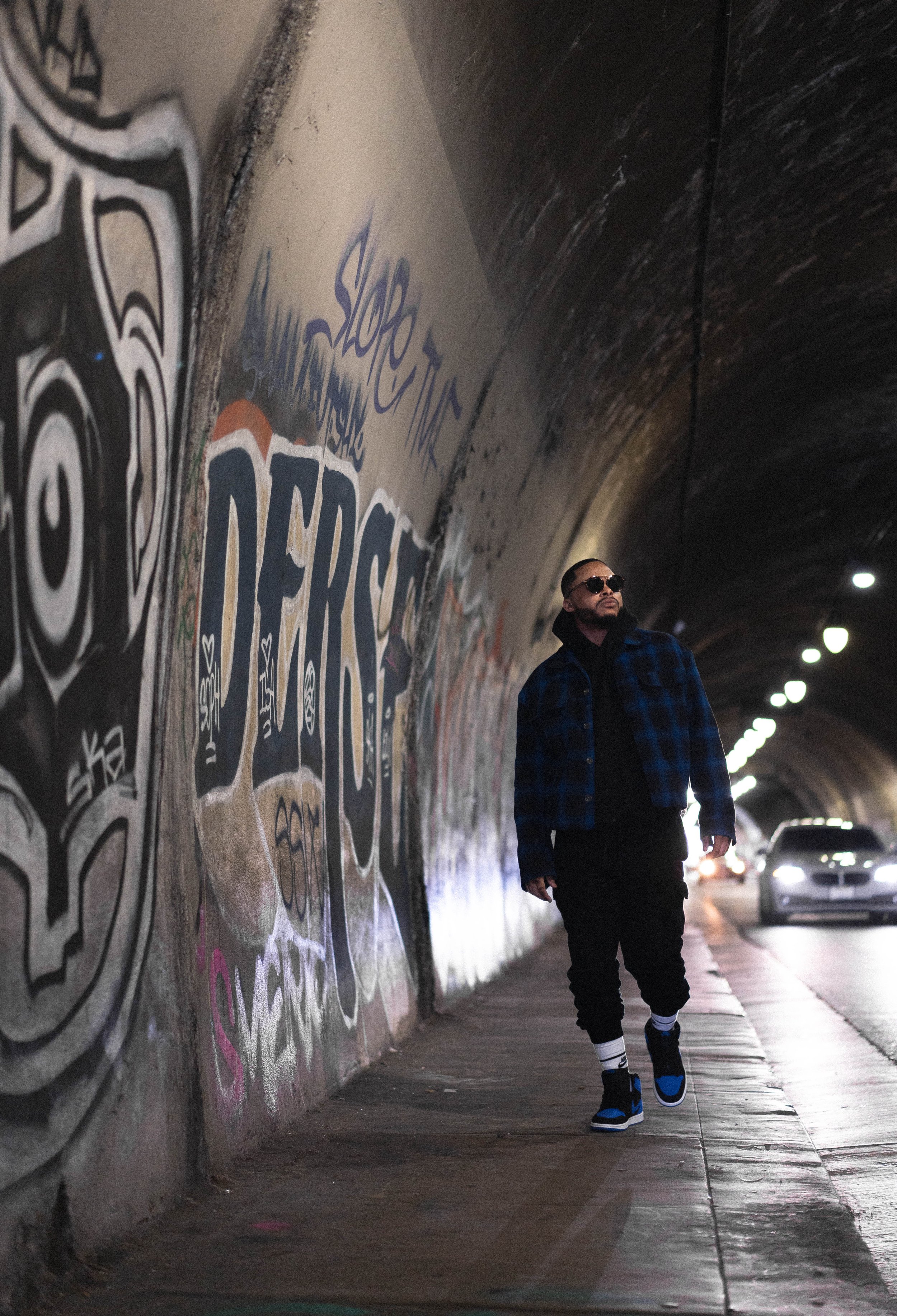 A man in a blue checkered jacket and sunglasses walking in a dark tunnel with graffiti on the wall and cars in the distance.
