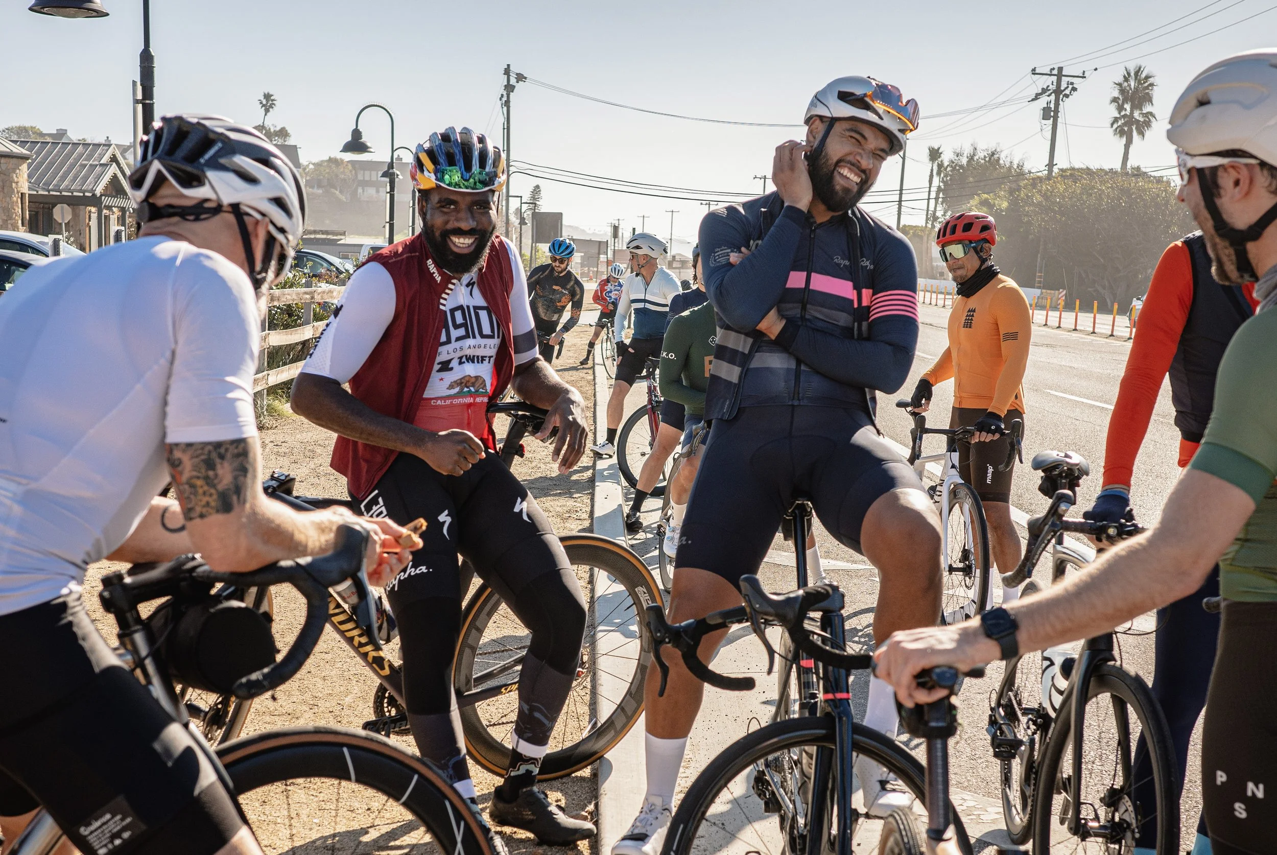 A group of cyclists wearing helmets and athletic clothing gathered on a sidewalk, smiling and talking, with some holding their bikes, in a sunny outdoor setting.