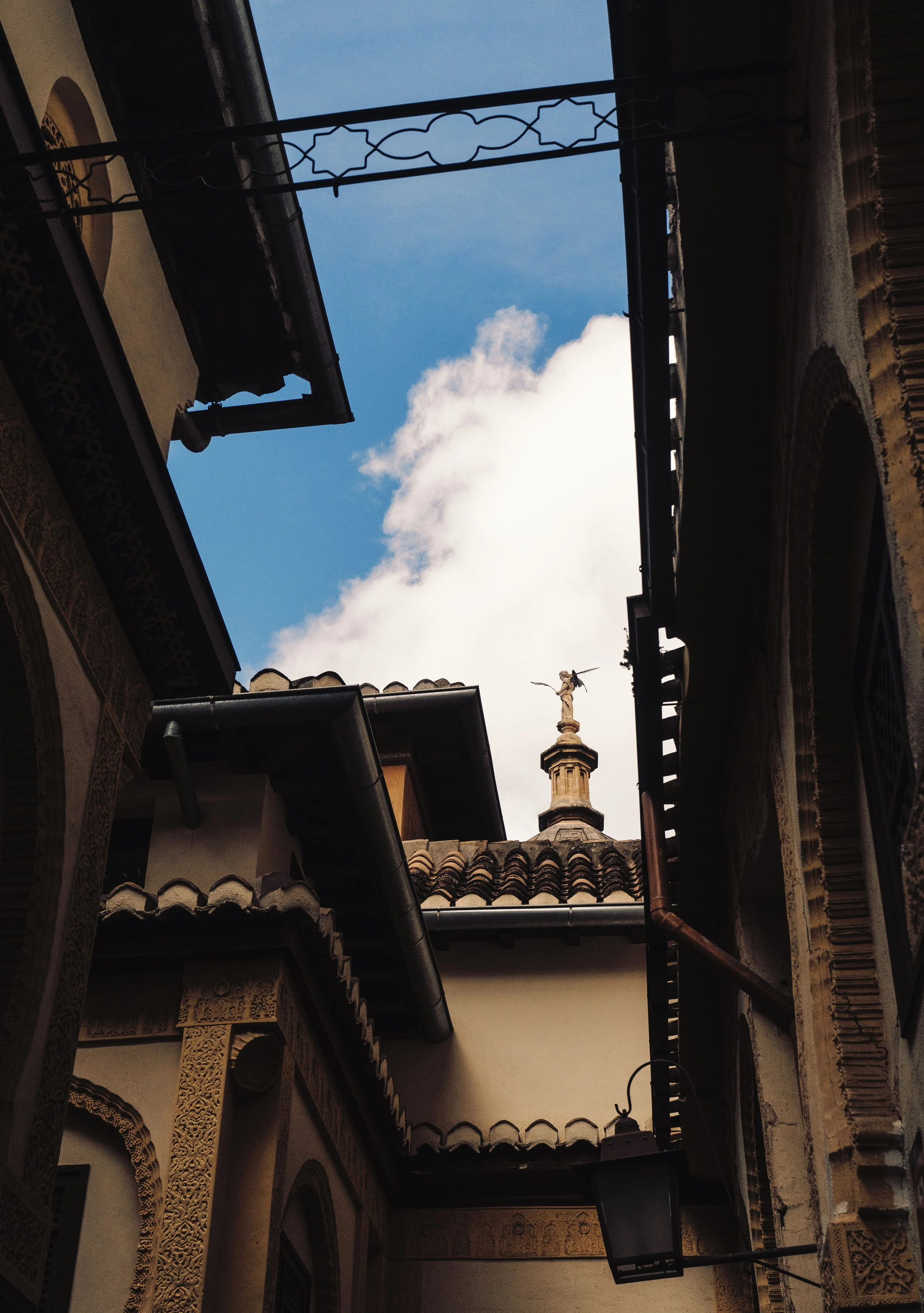 View of a courtyard with a building featuring ornate arches, textured stone details, a tiled roof, a rooftop cross, and a cloudy sky above.