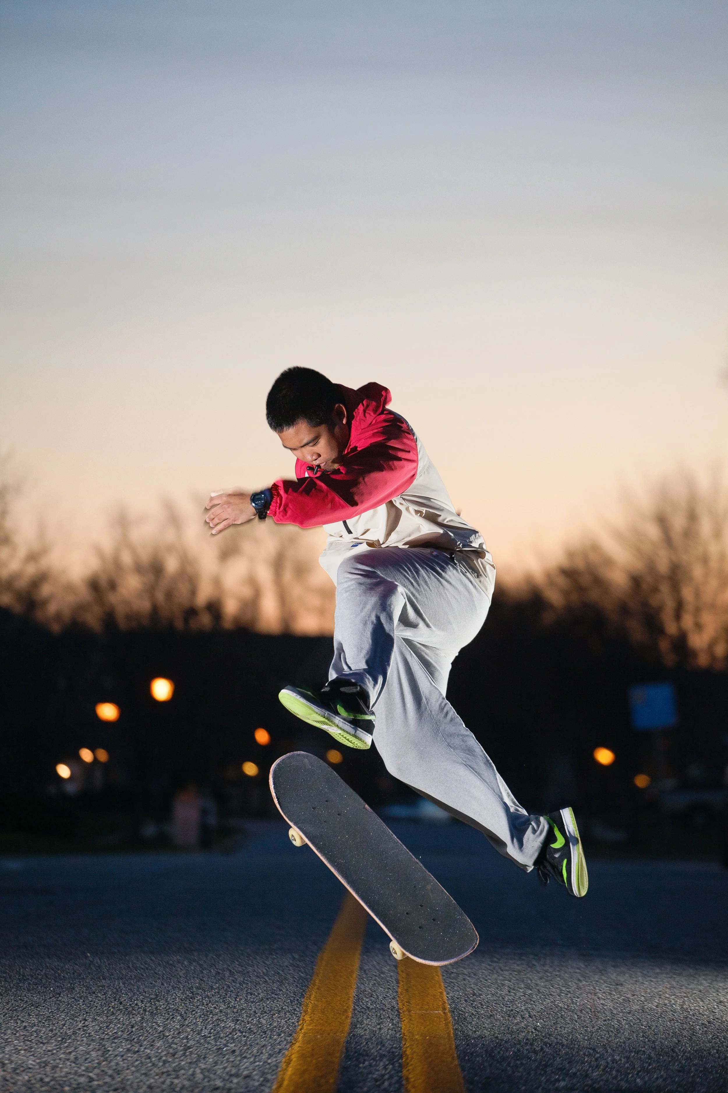 A person skateboarding on a road at sunset or dusk, wearing a red and white jacket, gray pants, and a black watch, performing a trick with the skateboard airborne. The background shows a darkening sky with some lights in the distance.