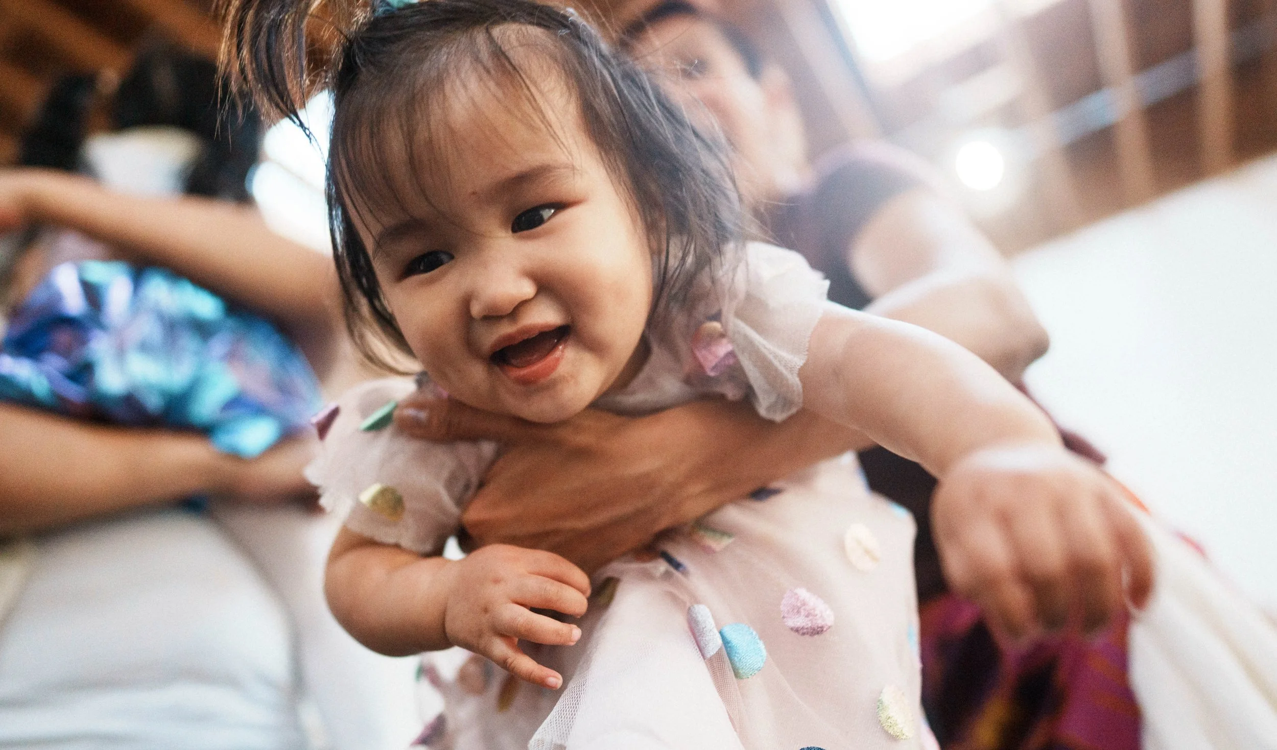 A young girl with dark hair lying on a person's arm, smiling with her mouth open, wearing a white dress with colorful polka dots.