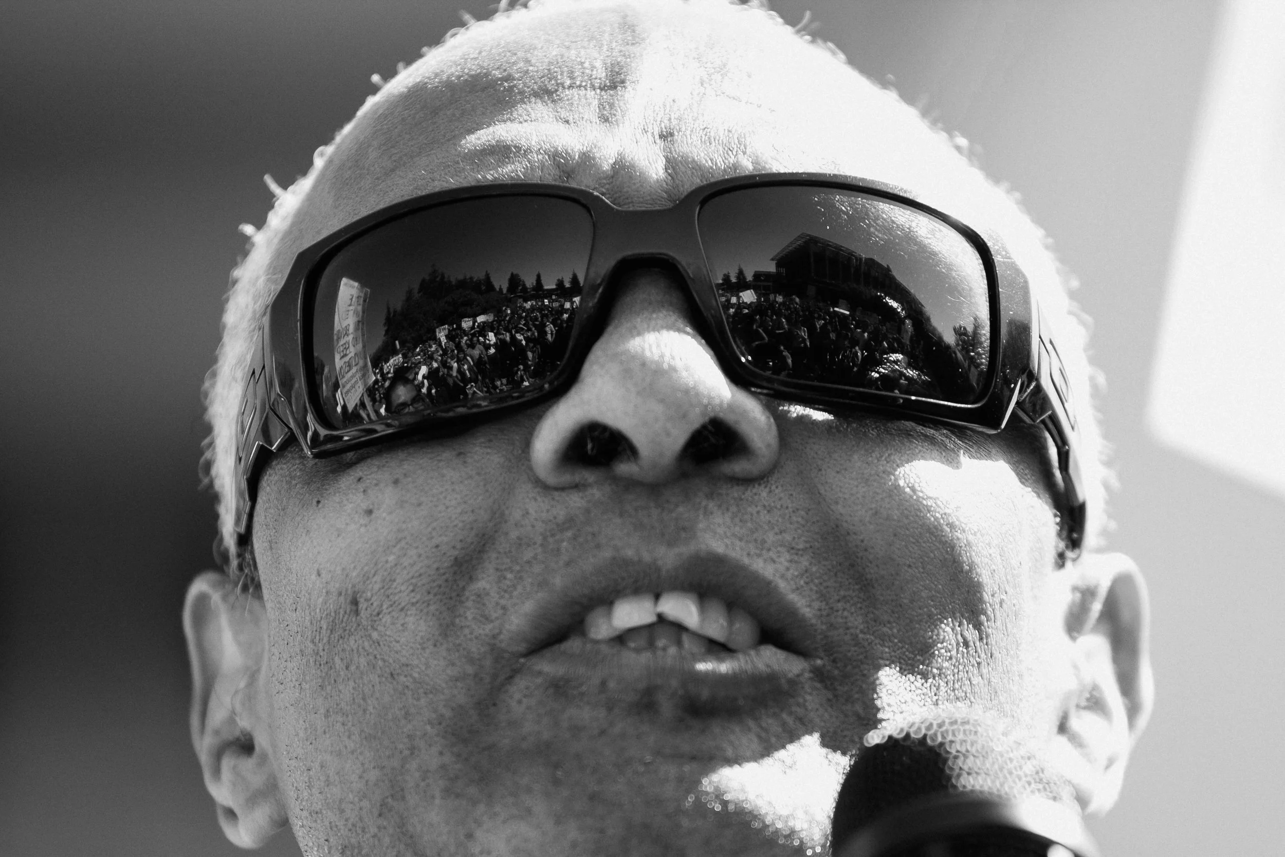 Black and white close-up photo of a person's face wearing sunglasses that reflect a crowd of people holding signs, with some trees and buildings in the background.