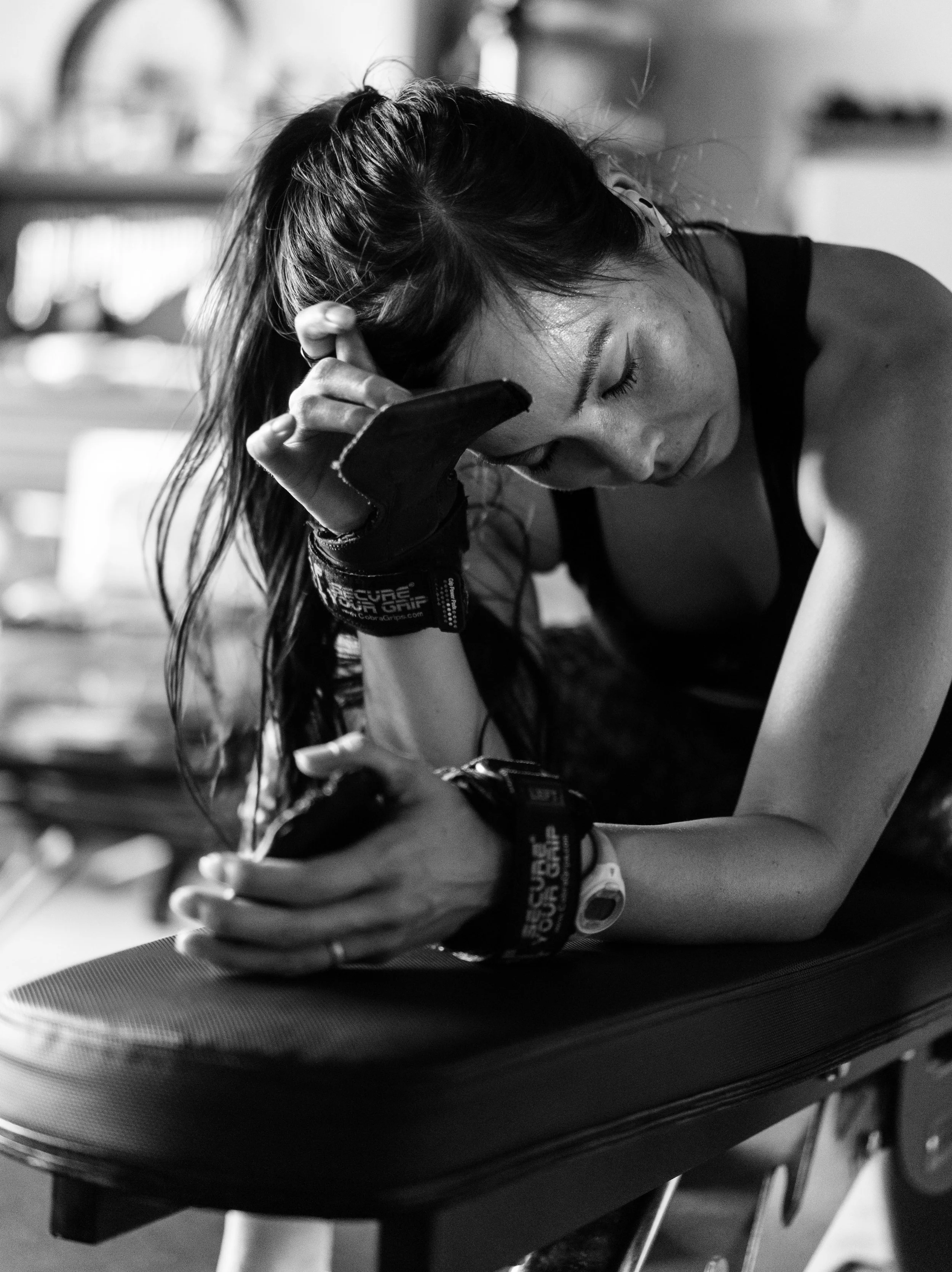 A woman in athletic gear resting her head on her arm amid exercise equipment, appearing exhausted or contemplative.
