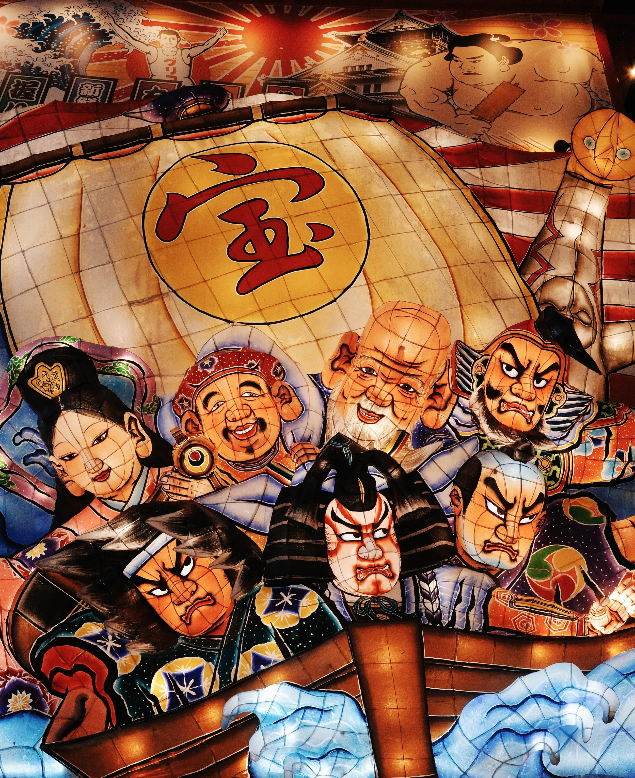 Colorful Japanese festival lanterns with traditional designs and characters, featuring a large smiling face, a stern face, and various decorative elements, illuminated in a vibrant display.