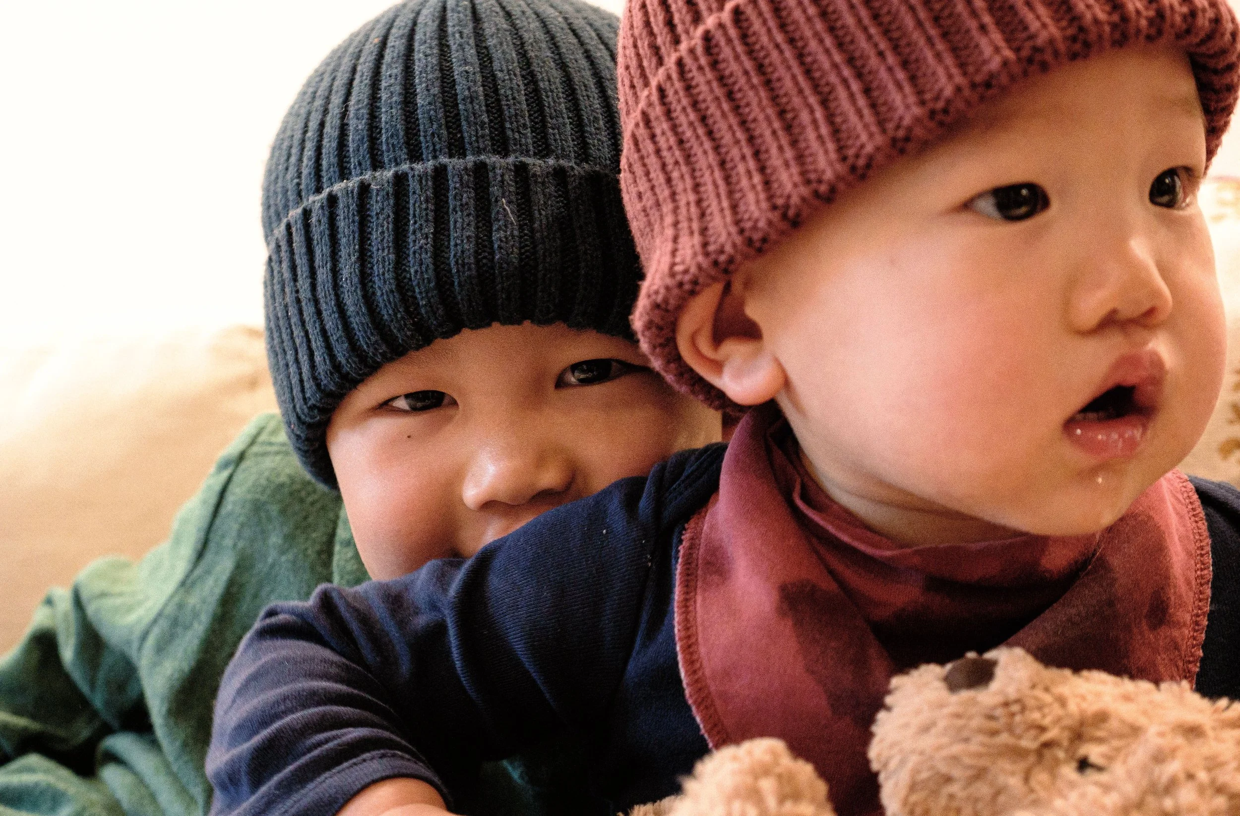Two young children wearing knit hats, one black and one maroon, are close together. They are indoors, leaning on a surface with a teddy bear partially visible in the foreground.