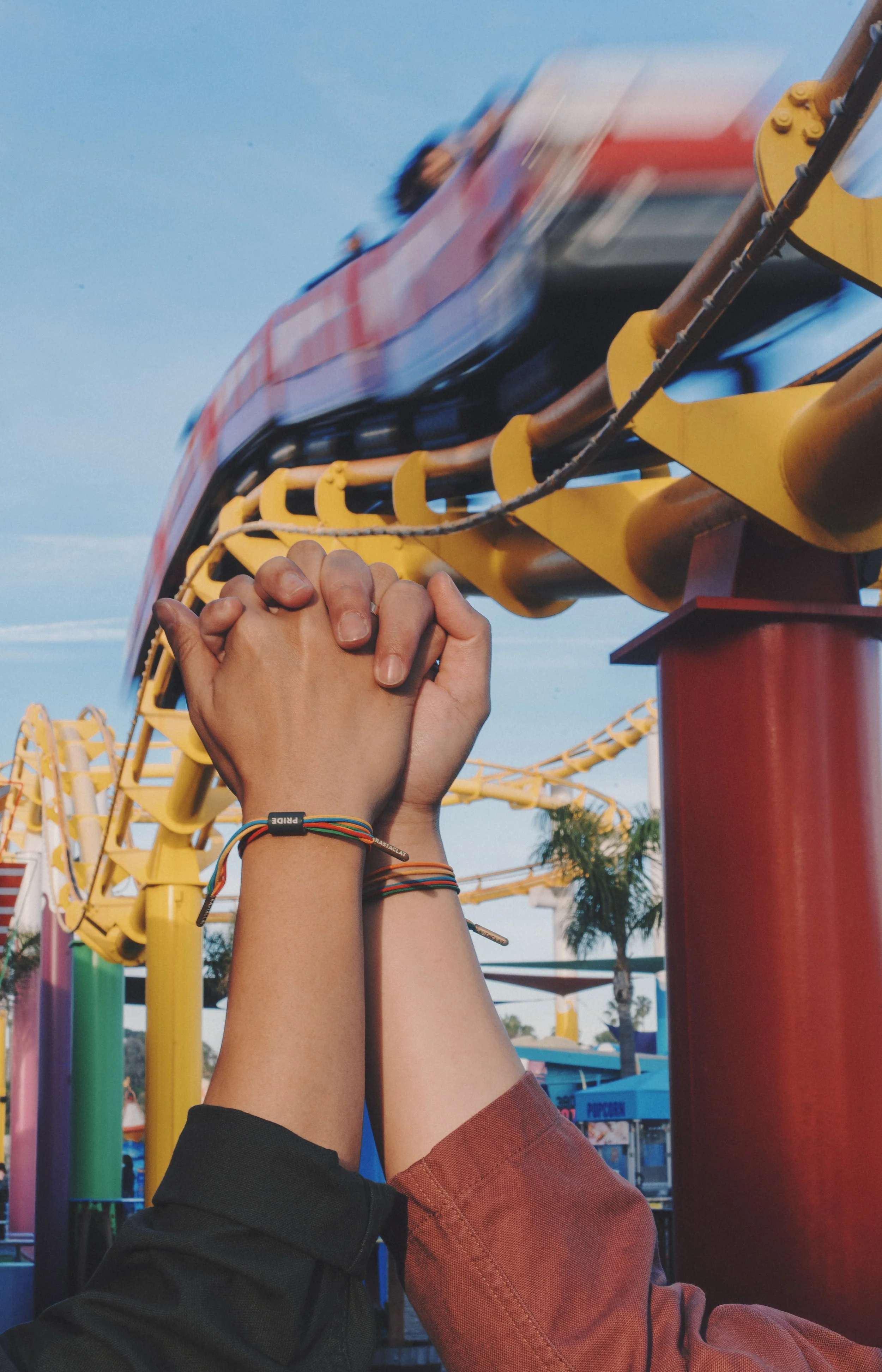 Two people holding hands in front of a roller coaster at an amusement park, with a blurred roller coaster and bright blue sky in the background.