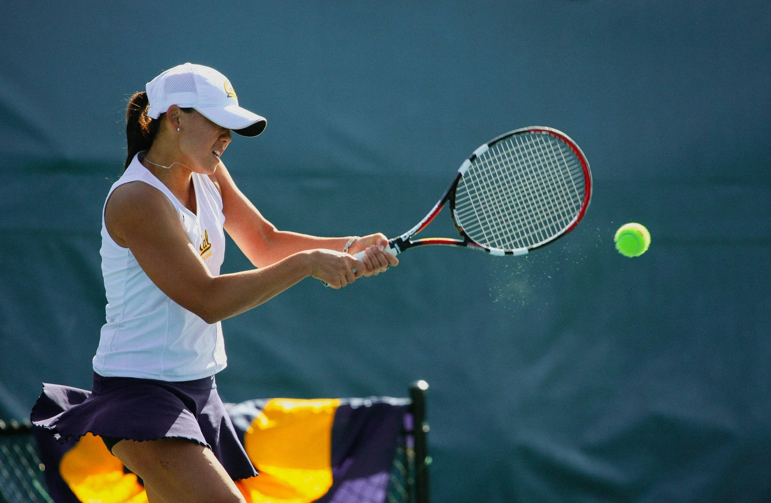 A female tennis player wearing a white cap and sleeveless shirt hitting a tennis ball with a racket on a court.