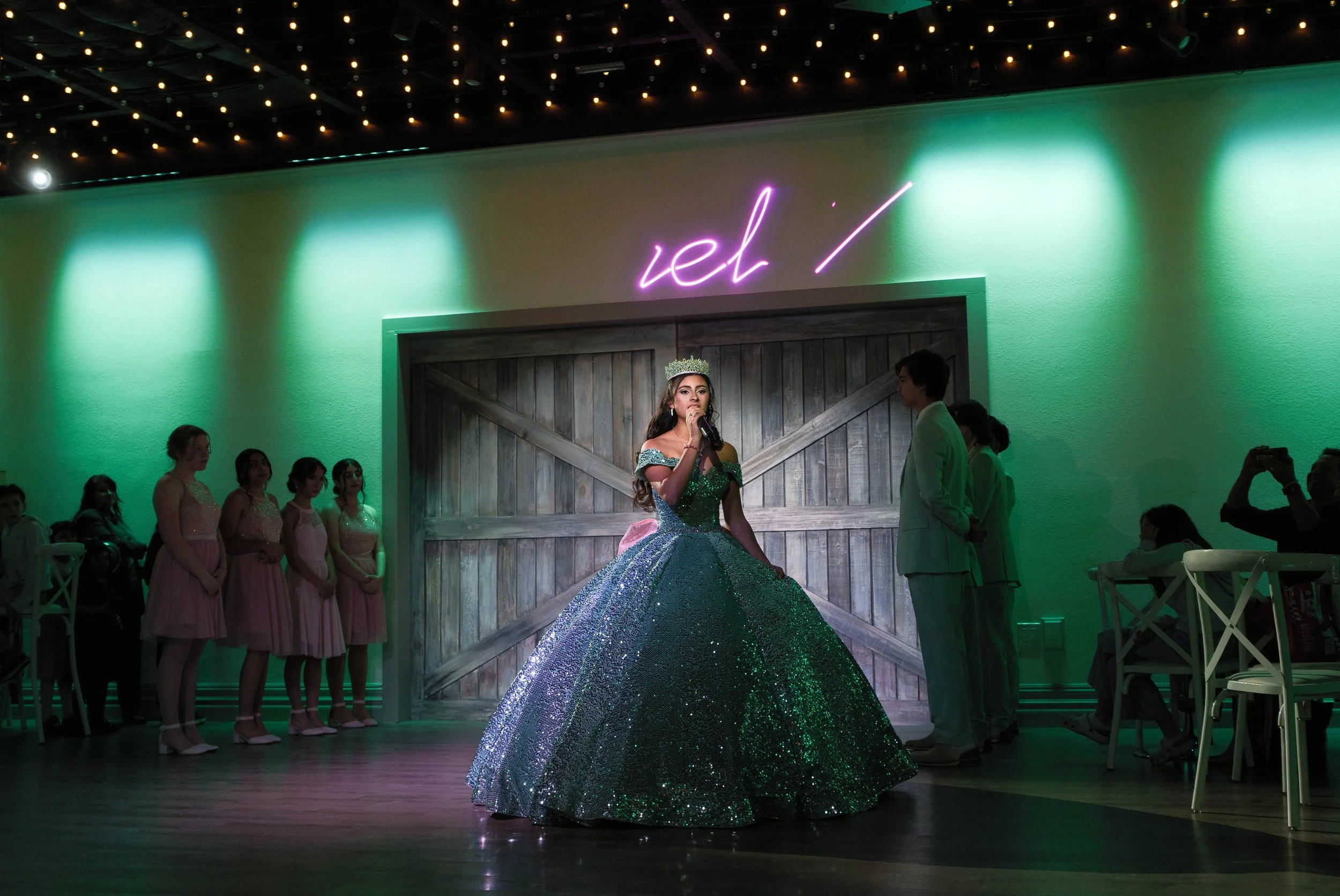 A girl dressed as a princess in a sparkling green gown and tiara performs on stage with a microphone, surrounded by young girls in pink dresses and a few adults, in a decorated indoor setting with string lights and a neon sign reading 'ieh/' on the w
