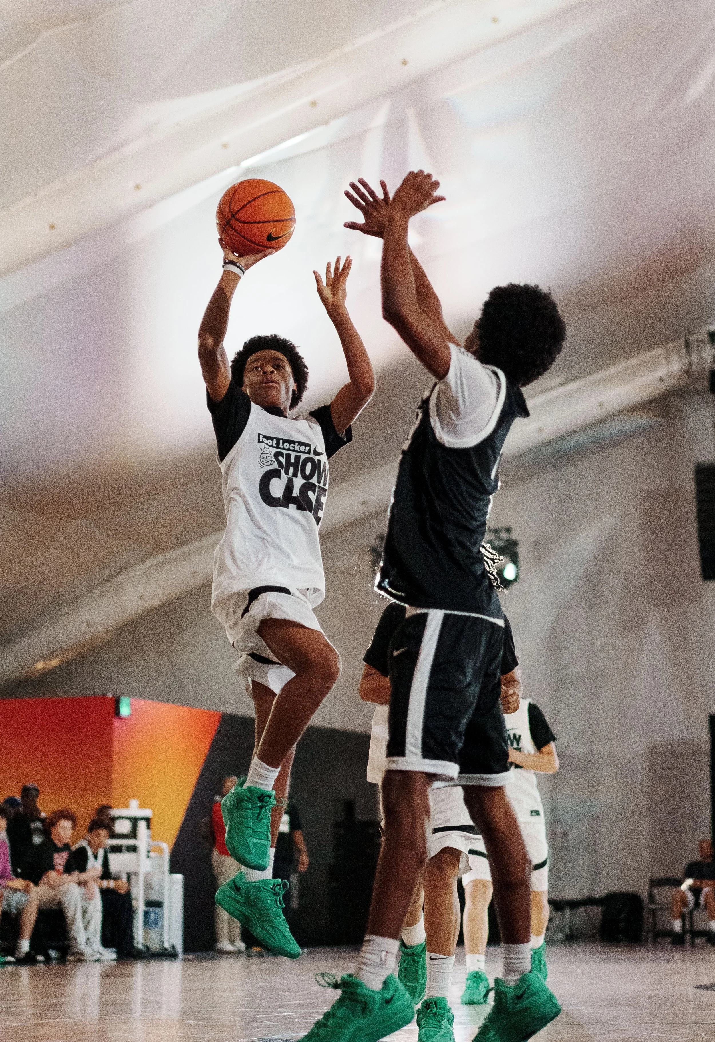 A basketball game in an indoor court with players in black and white uniforms. One player is jumping to make a shot while another is attempting to block. The audience and bench are visible in the background.