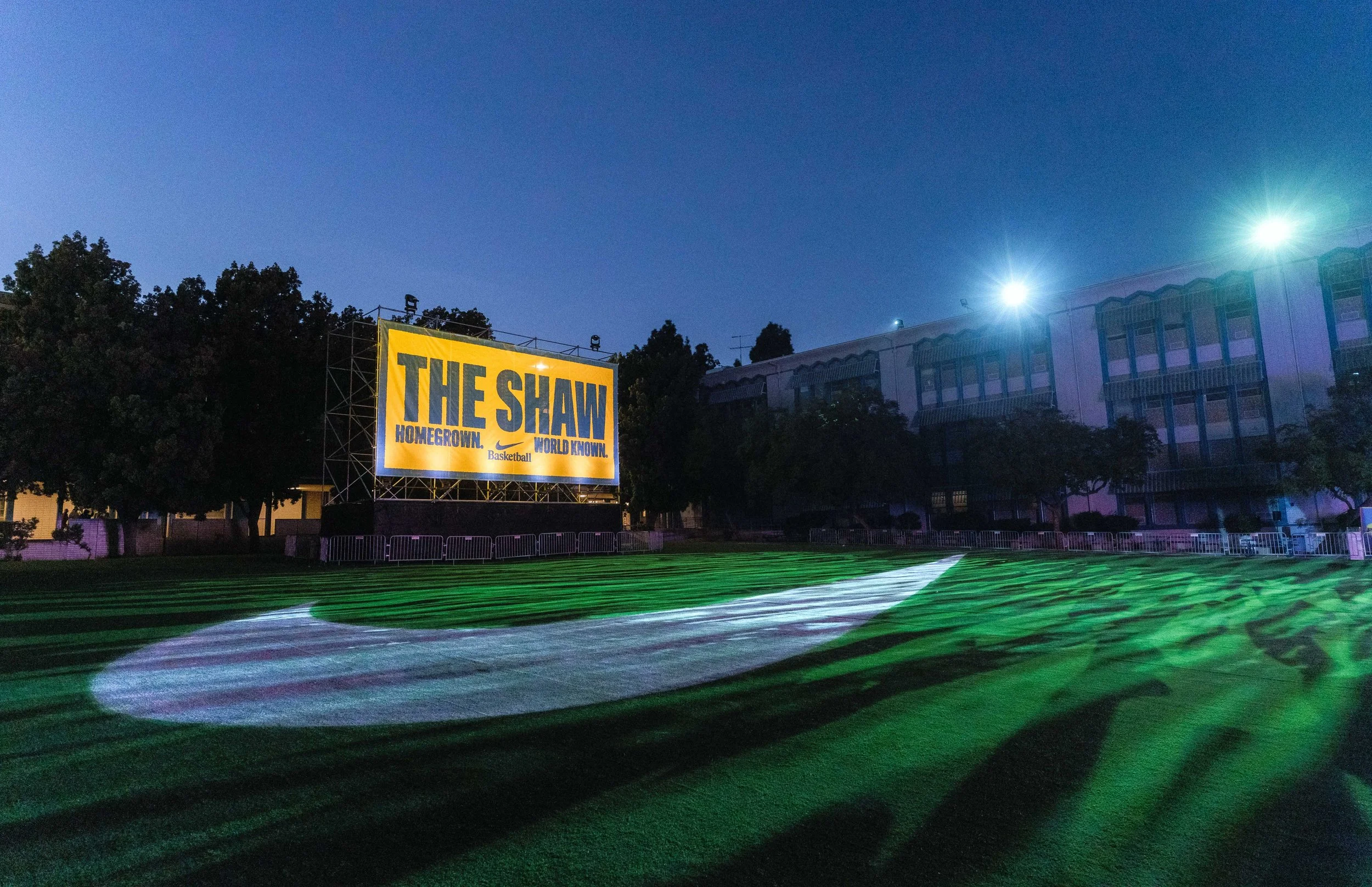 Empty football field at dusk with a large yellow sign that reads 'THE SHAW HOMEGROWN. WORLD KNOWN.' and bright stadium lights illuminating the scene.