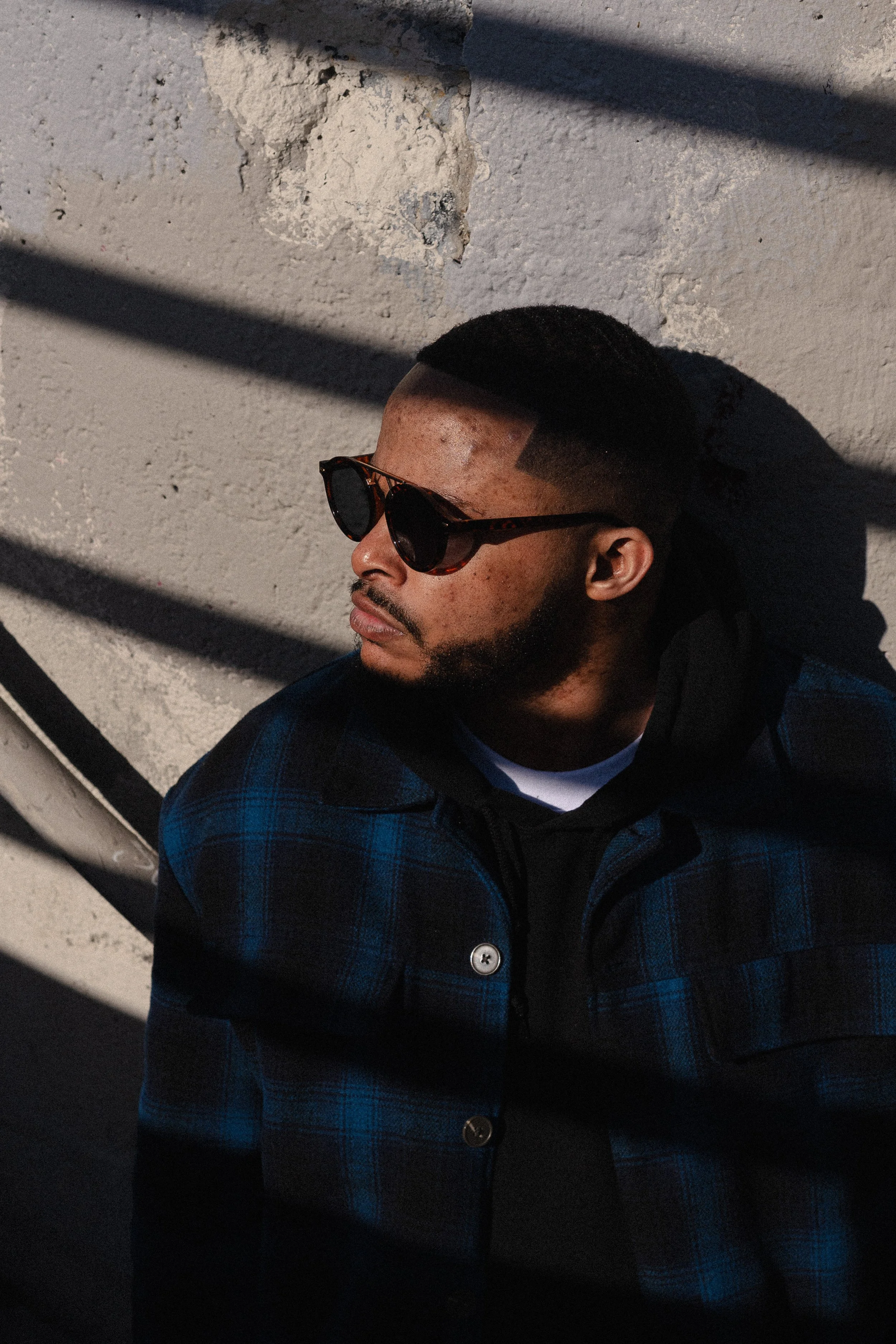 A man with a close-cropped haircut wearing sunglasses and a blue plaid jacket, leaning against a textured, light-colored wall with shadows cast across it.