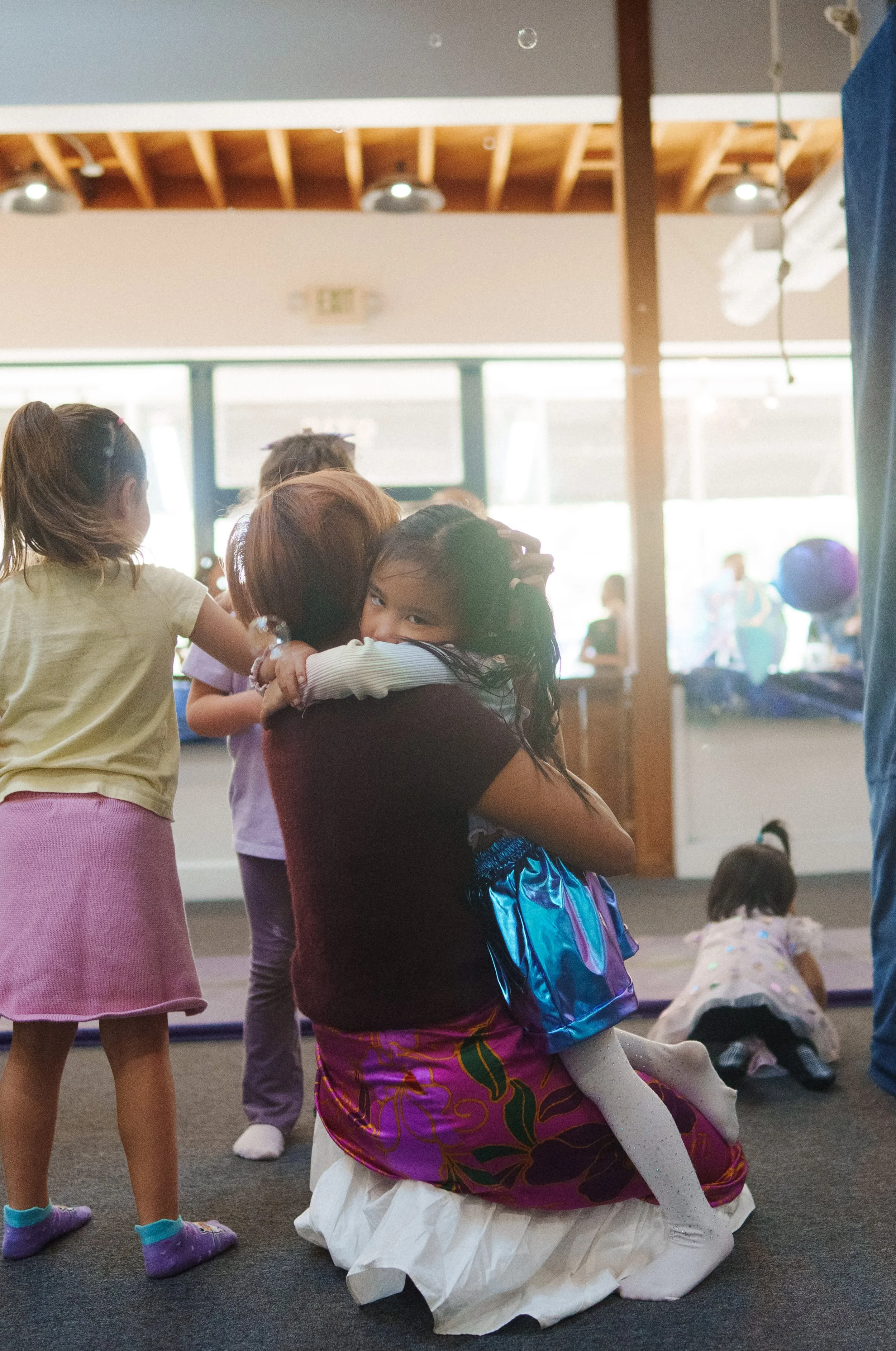 A woman in a colorful skirt and white blouse is kneeling on the floor, hugging a young girl on her lap. The girl is wearing a blue shiny dress, white glittery tights, and has a backpack. Other children are nearby, some standing and some sitting or ly