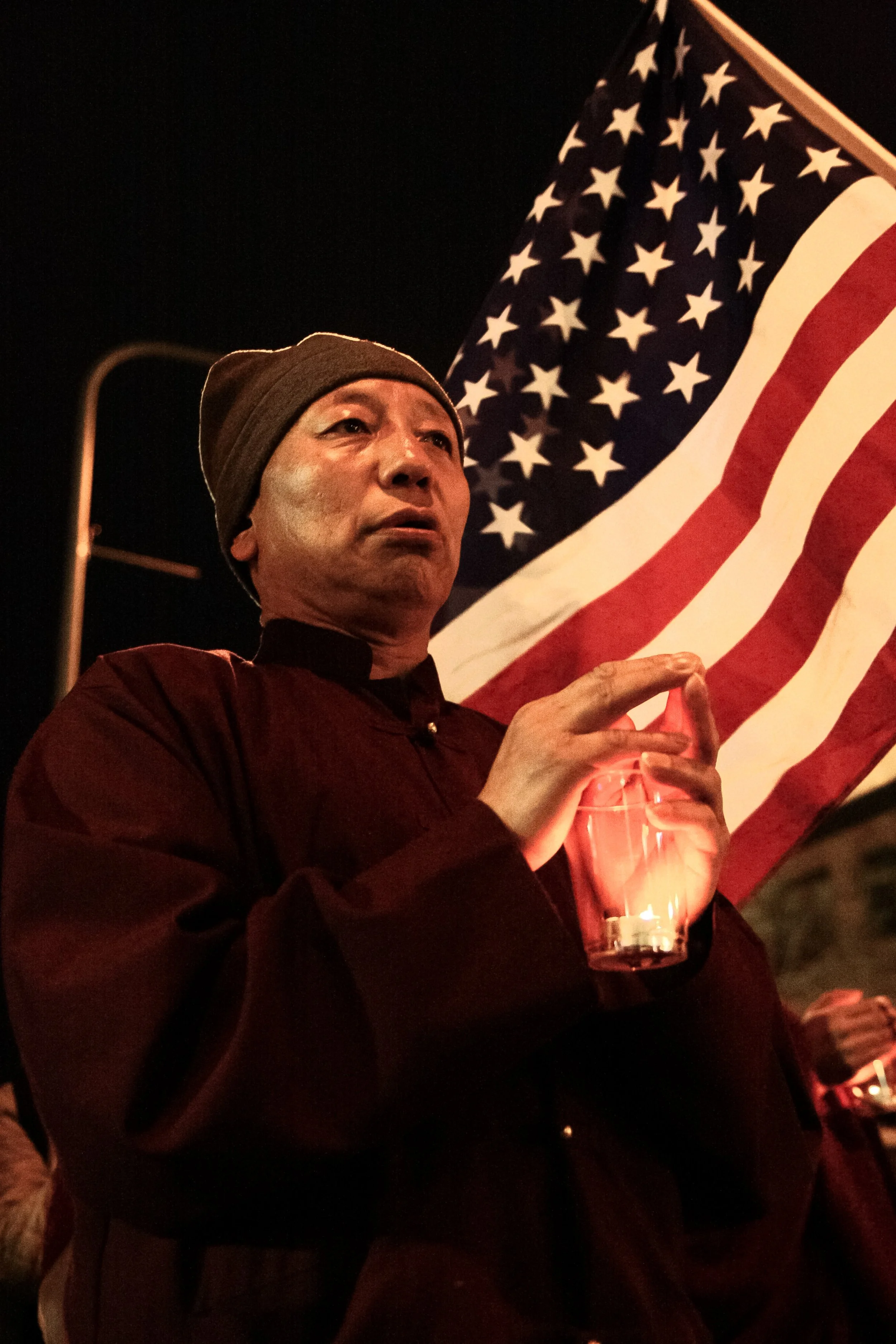A man in a brown beanie holding a candle in a glass container, standing in front of an American flag at night.