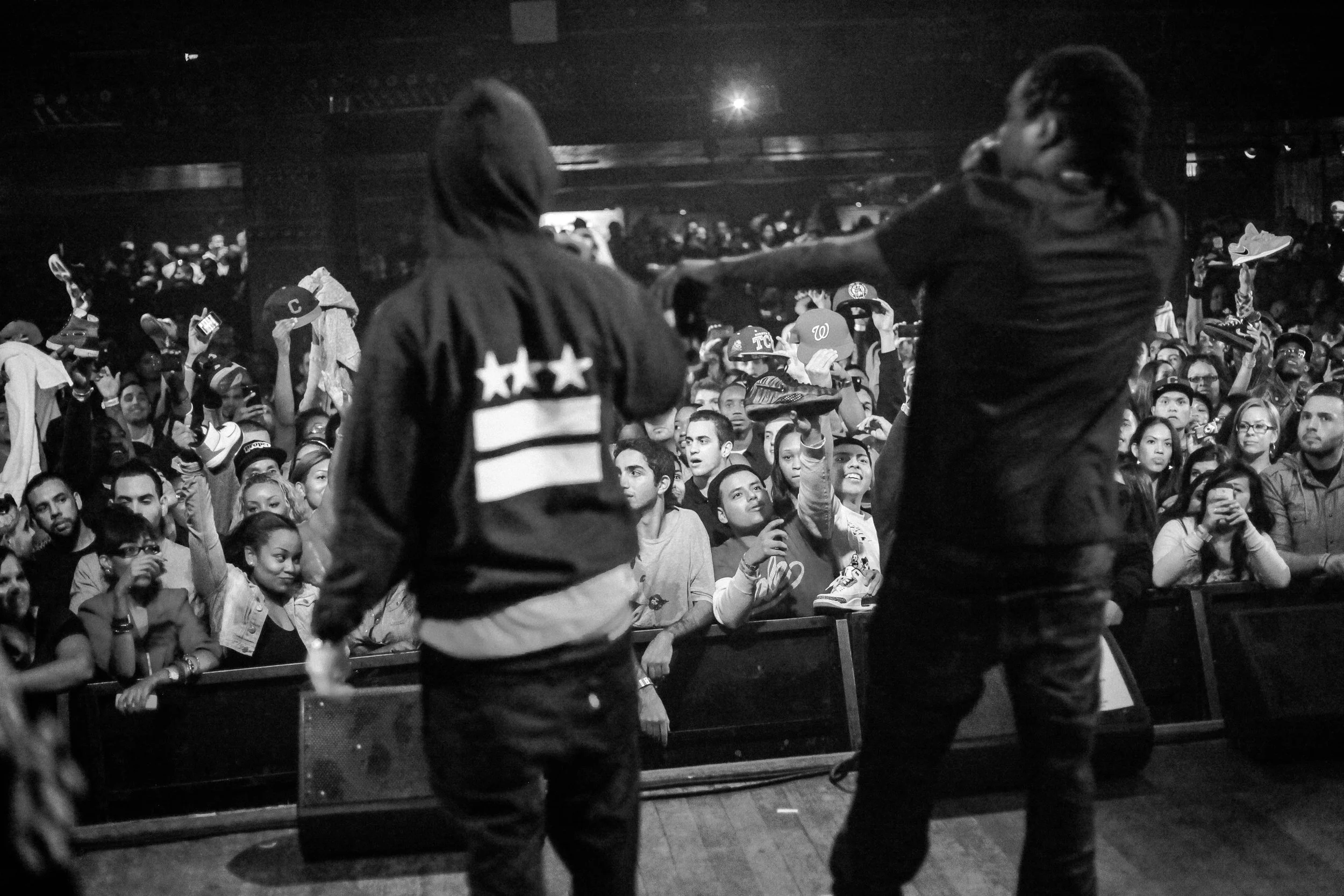 Crowd of people at a concert, with two security guards standing in the foreground. Many people in the crowd are holding their phones or hats, and some are reaching out toward the stage.