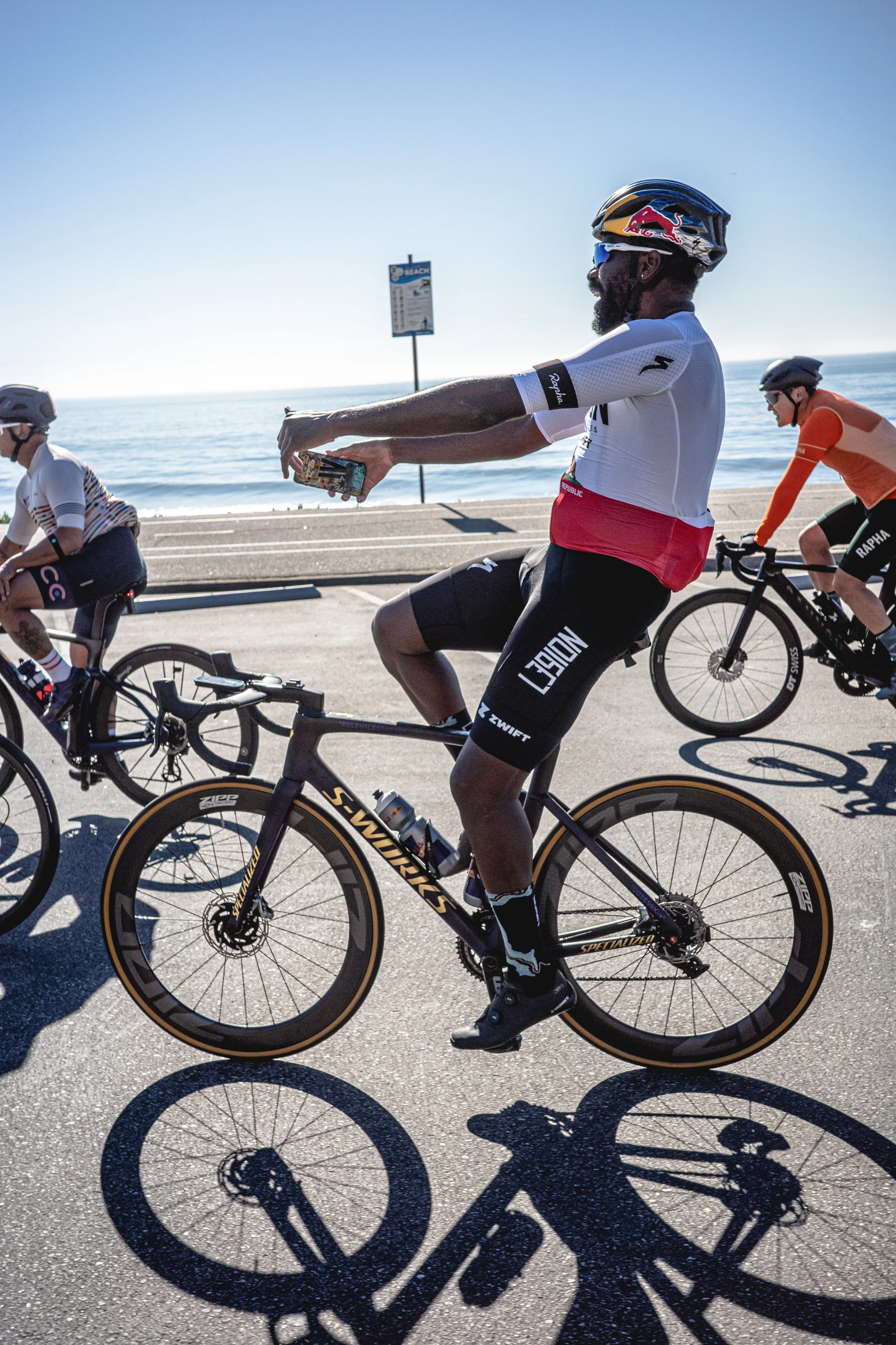 Cyclists riding along a beachside road with their shadows cast on the pavement, at a sunny beach location.