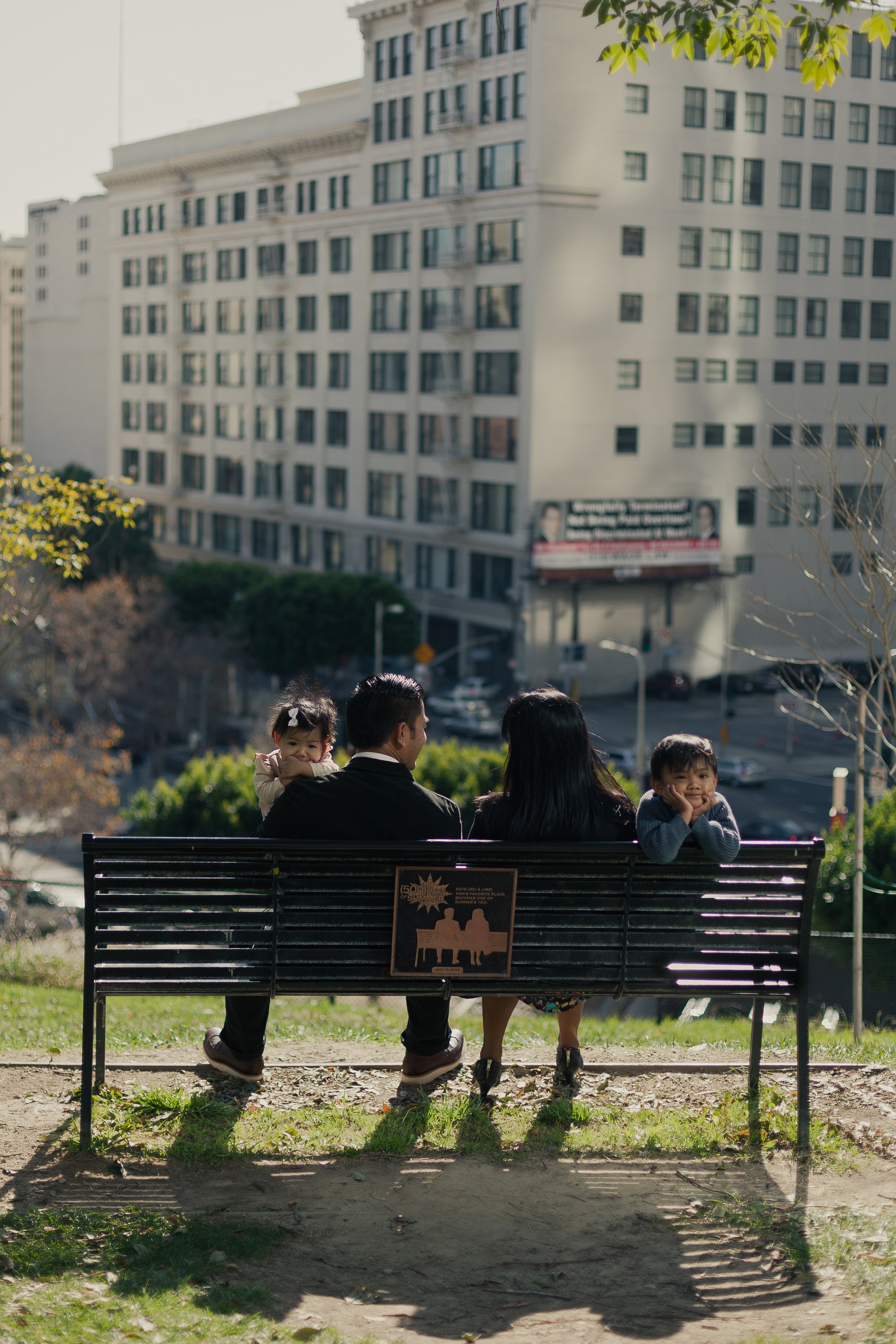 Four people sitting on a park bench in an urban park, with two children and two adults. The children are facing forward, with one girl resting her head on an adult's shoulder and a boy smiling with hands on his face. The adults are facing away from t