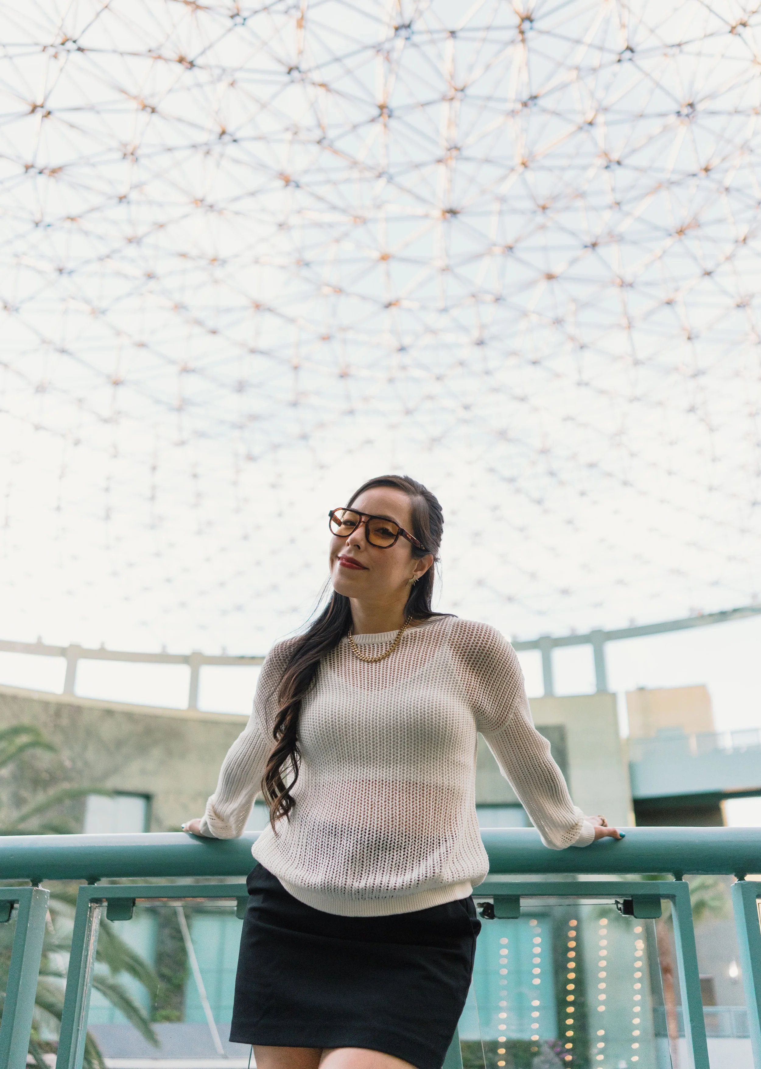 A woman wearing glasses, a cream sweater, and a black skirt standing outdoors in front of a modern architectural structure with a large, glass-domed roof, posing with her hands on a railing.