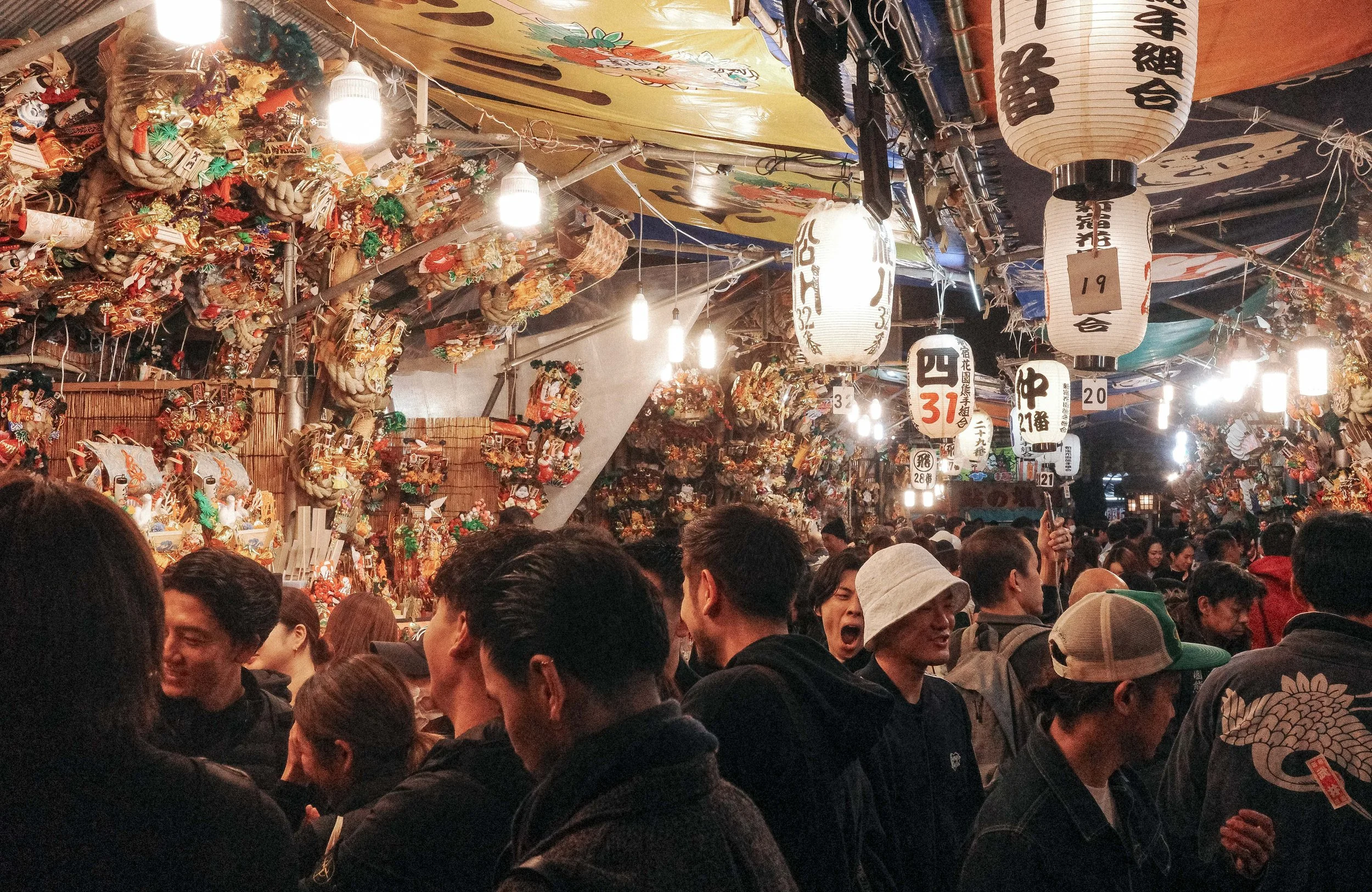 A crowded marketplace or festival scene in Japan with hanging paper lanterns and various decorative ornaments, with many people in the foreground.