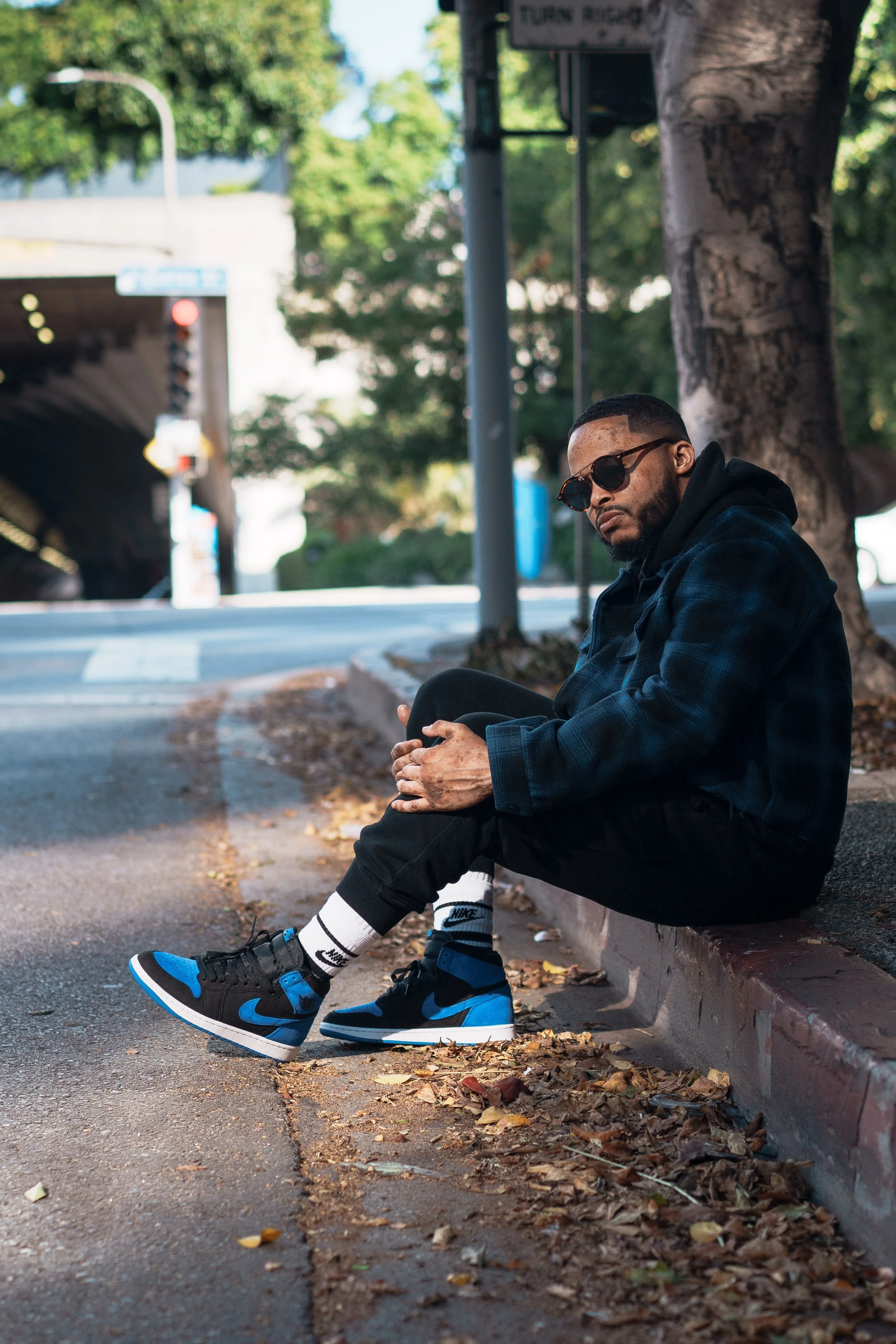 A man sitting on a curb under a tree, wearing sunglasses, a black hoodie, a dark jacket, black pants, and blue and black Nike sneakers. The scene appears to be outdoors in a city or suburban area, with a street, trees, and a bridge or tunnel in the b