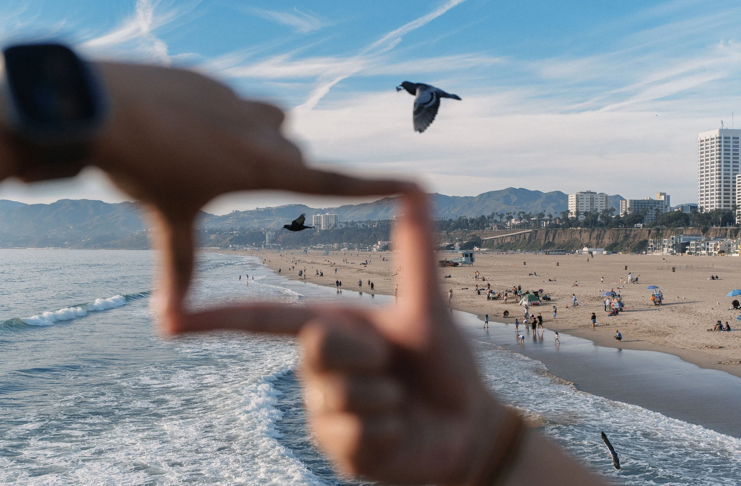 A beach scene seen through the blurred hand of a person holding a camera. There are people on the sandy beach and in the water, with some umbrellas and tents, and high-rise buildings and mountains in the background. A bird is flying in the sky.