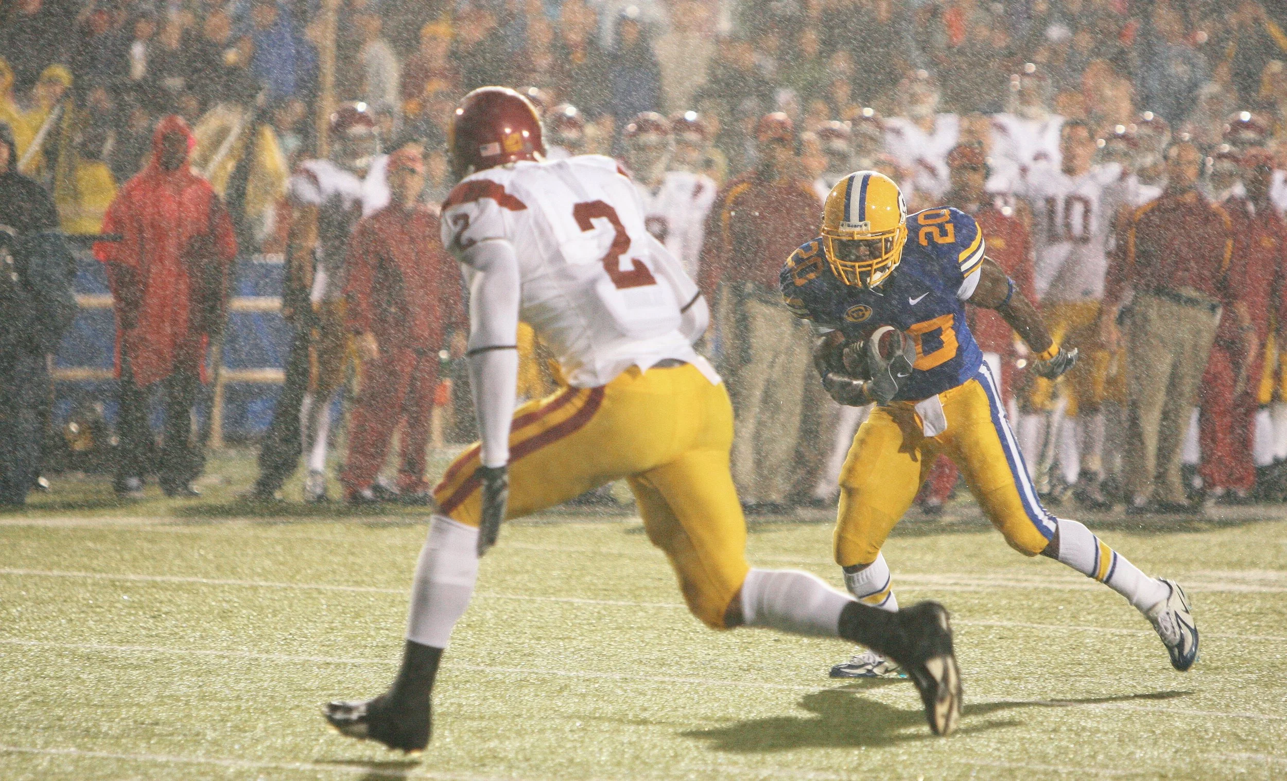 An American football game between USC and California taking place in the rain, with players in uniforms running on the field and spectators watching from the sidelines.