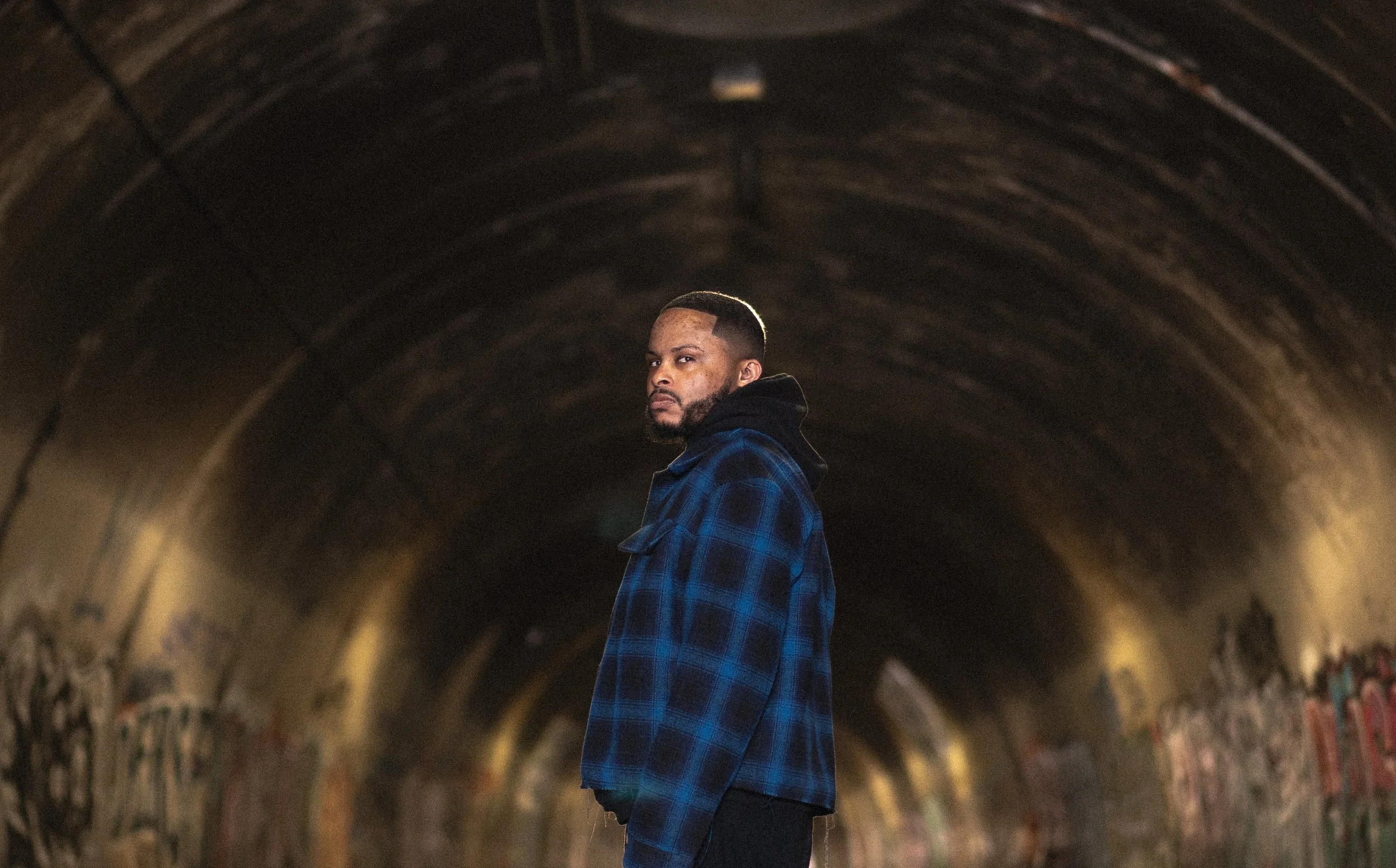 A man stands inside a dark, graffiti-covered tunnel with graffiti on the walls and curved concrete ceiling.