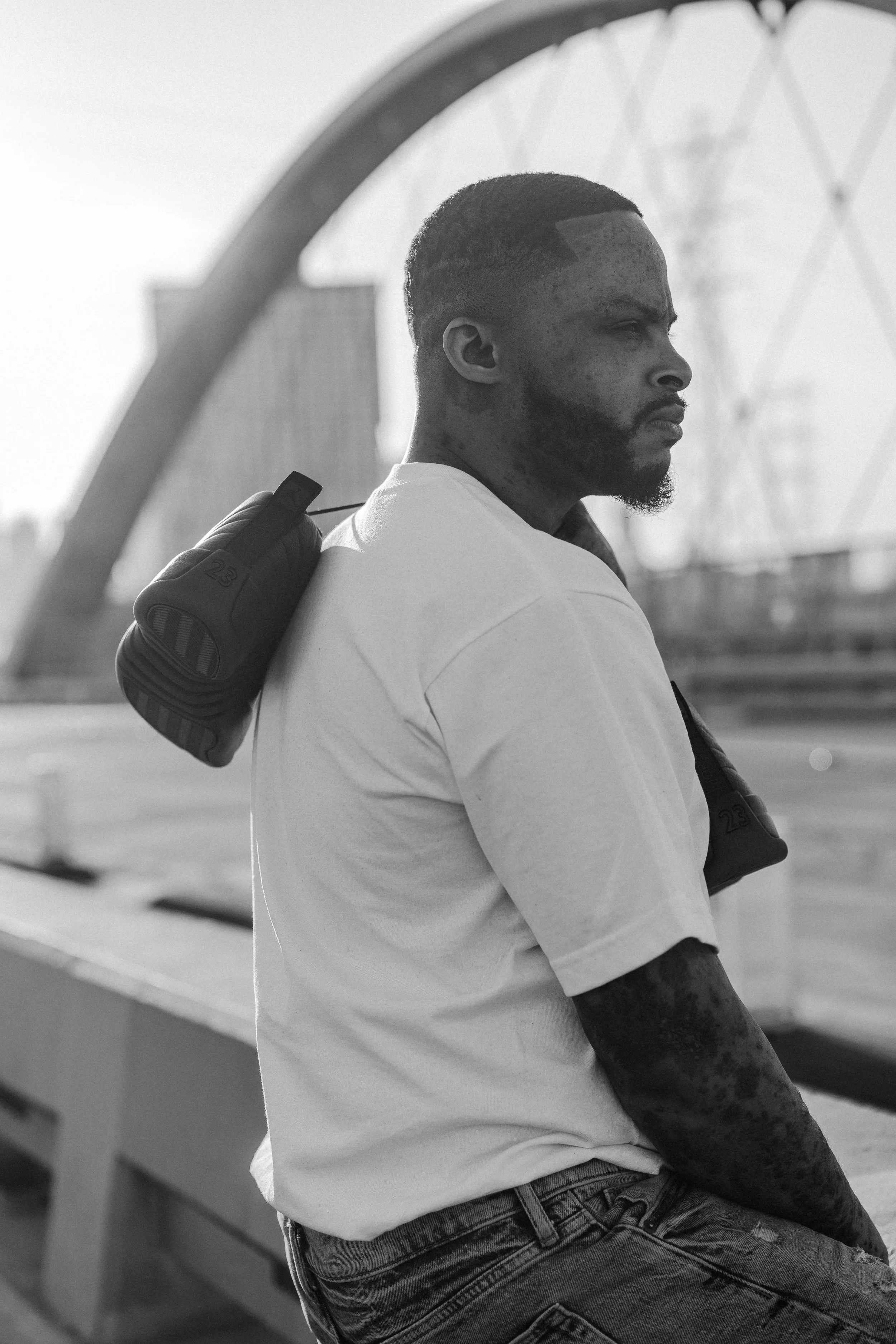 A man with a beard and short hair, wearing a white T-shirt and jeans, is leaning against a ledge or railing with his arms crossed. There is a large Ferris wheel in the background, indicating an outdoor setting.