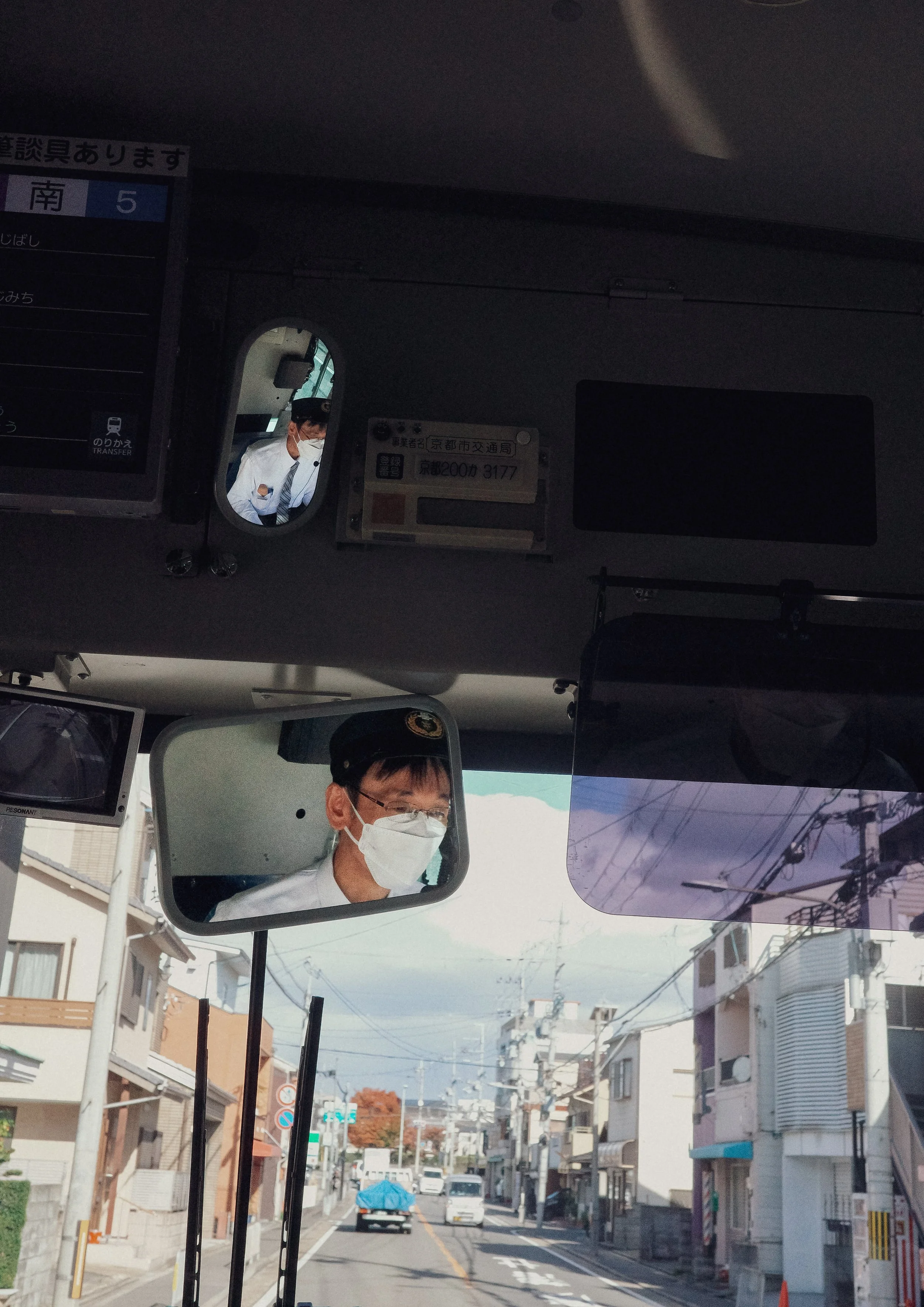 View of a street seen from inside a vehicle, with multiple mirrors showing the driver and part of the road behind, including cars, buildings, power lines, and a street sign.